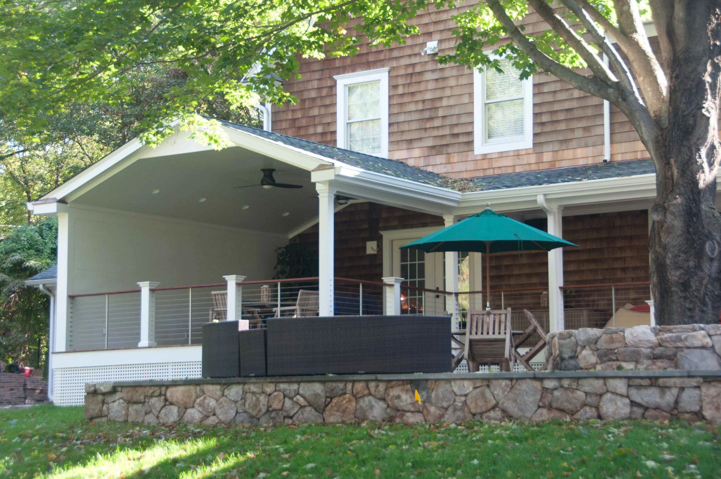 Backyard patio with furniture, umbrella, and trees in front of a house with brown siding and white trim.
