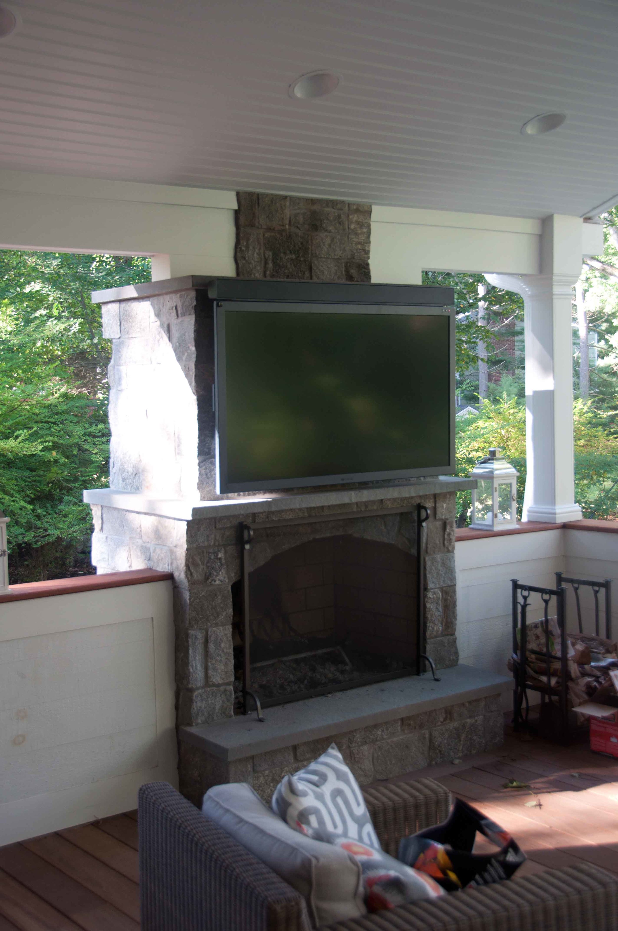 Outdoor porch area with stone fireplace and mounted flat-screen TV, surrounded by white walls and greenery.