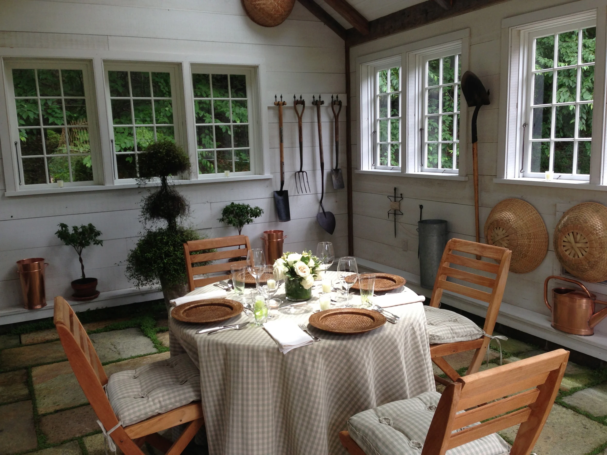 Dining area in a garden room with wooden chairs, a round table with a checkered tablecloth, a floral centerpiece, glassware, and place settings. The room has white paneled walls, multiple windows, gardening tools hanging on the wall, and decorative w