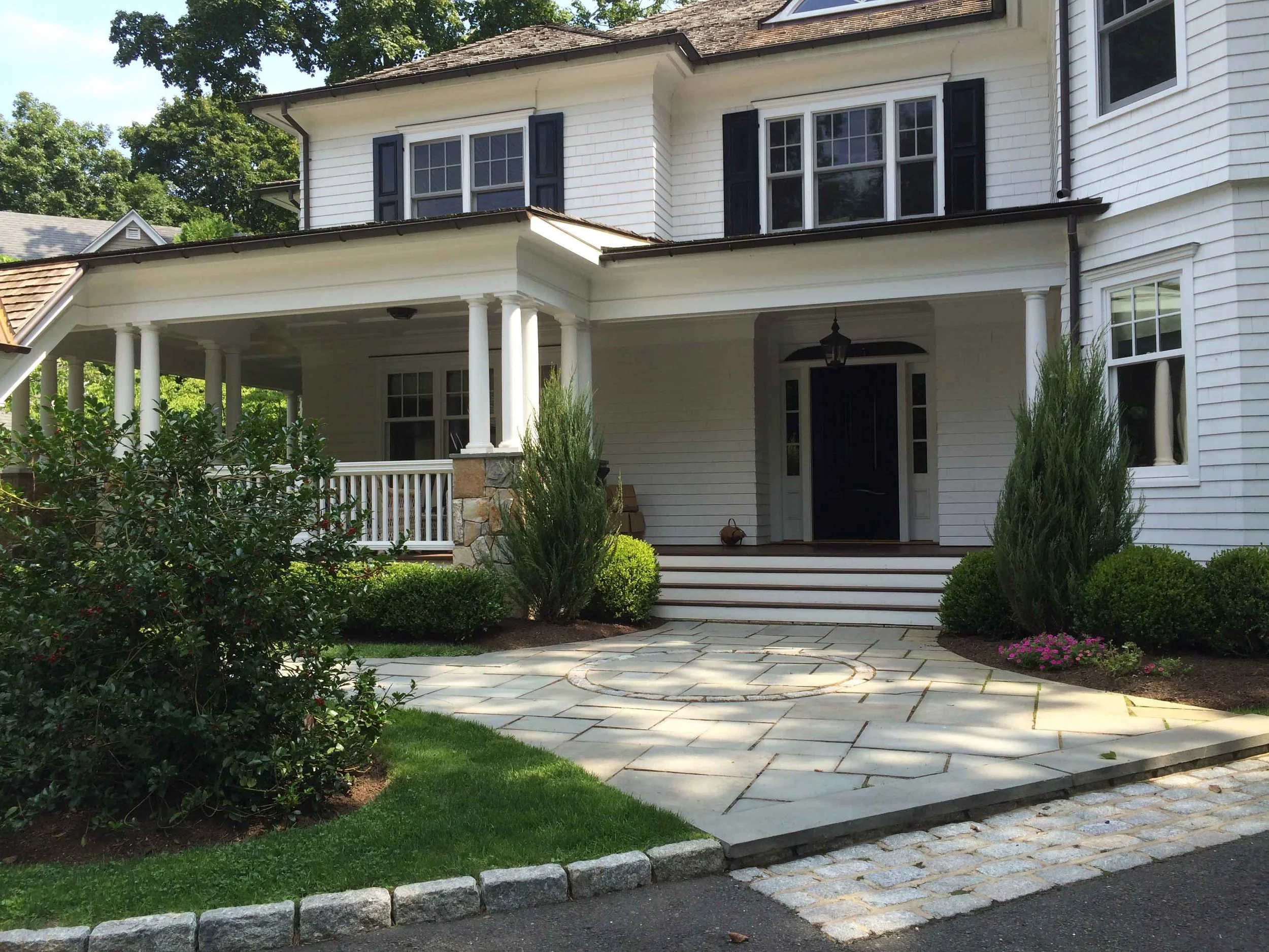 Front view of a white two-story house with a porch, black front door, and landscaped yard with bushes, small trees, and a stone walkway.