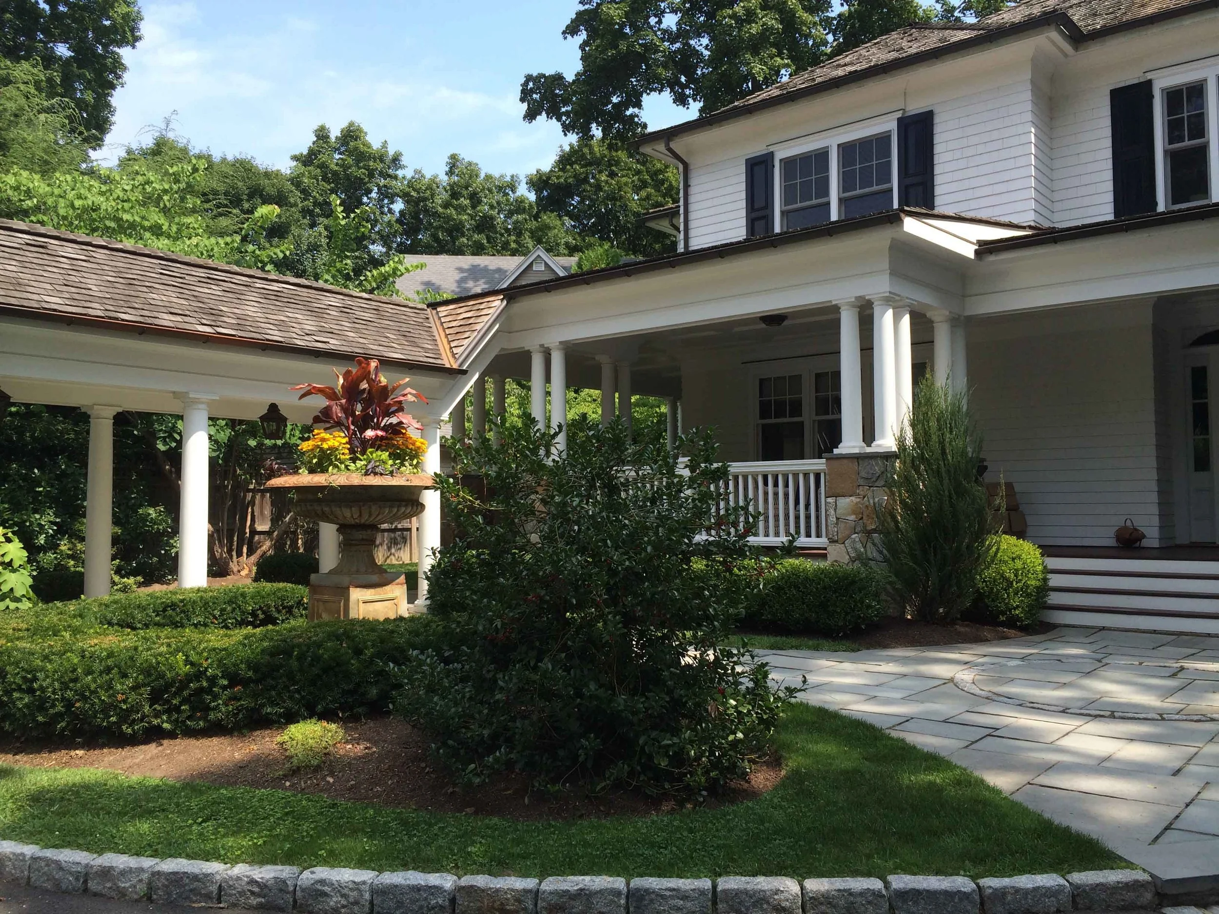Front yard of a house with a stone patio, green bushes, a flower planter, and a white house with columns and dark shutters.