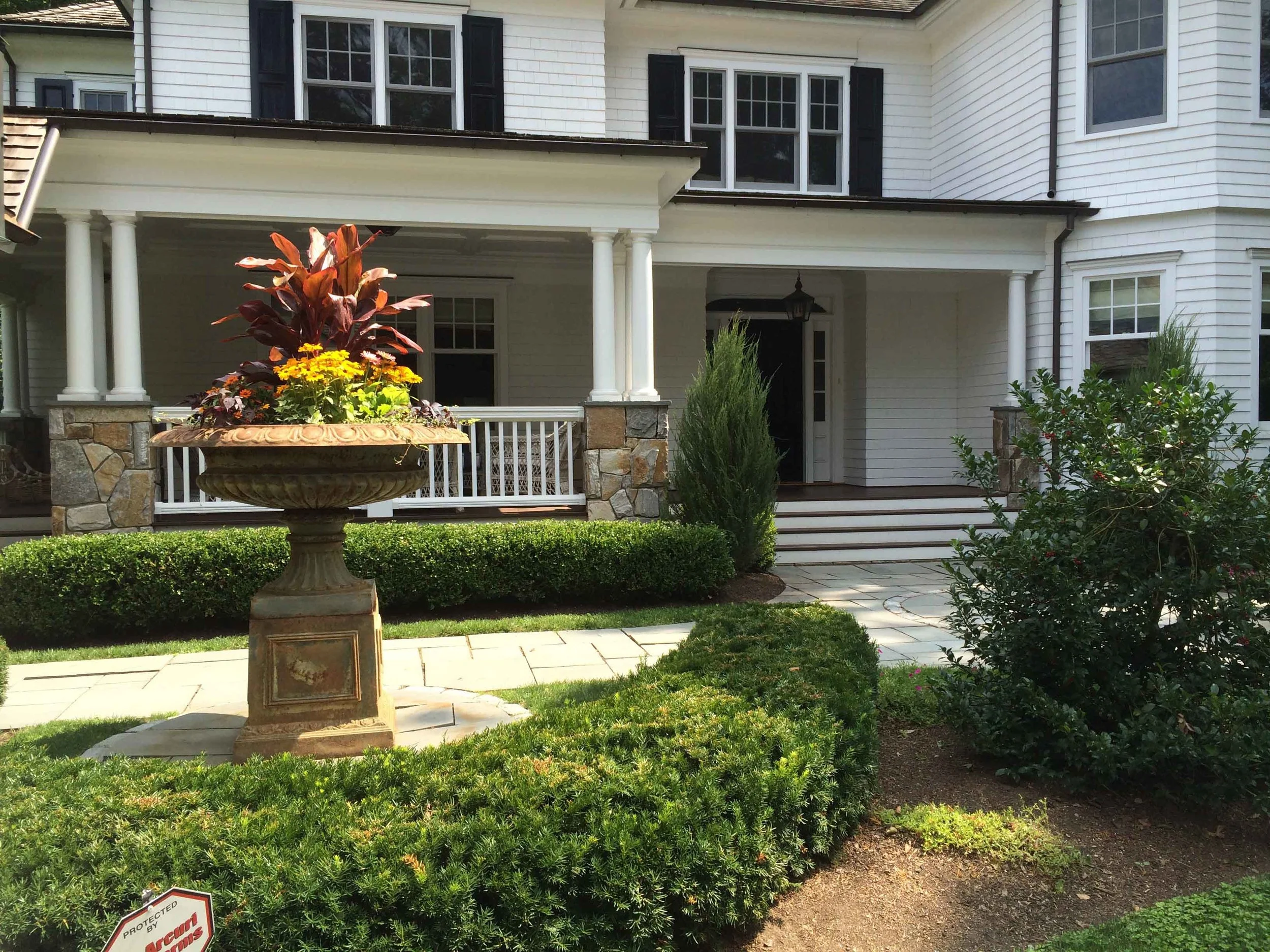 Front view of a large white house with black shutters, a porch with white railings and stone pillars, and a garden with a decorative stone planter filled with red and yellow flowers, bushes, and a small tree.