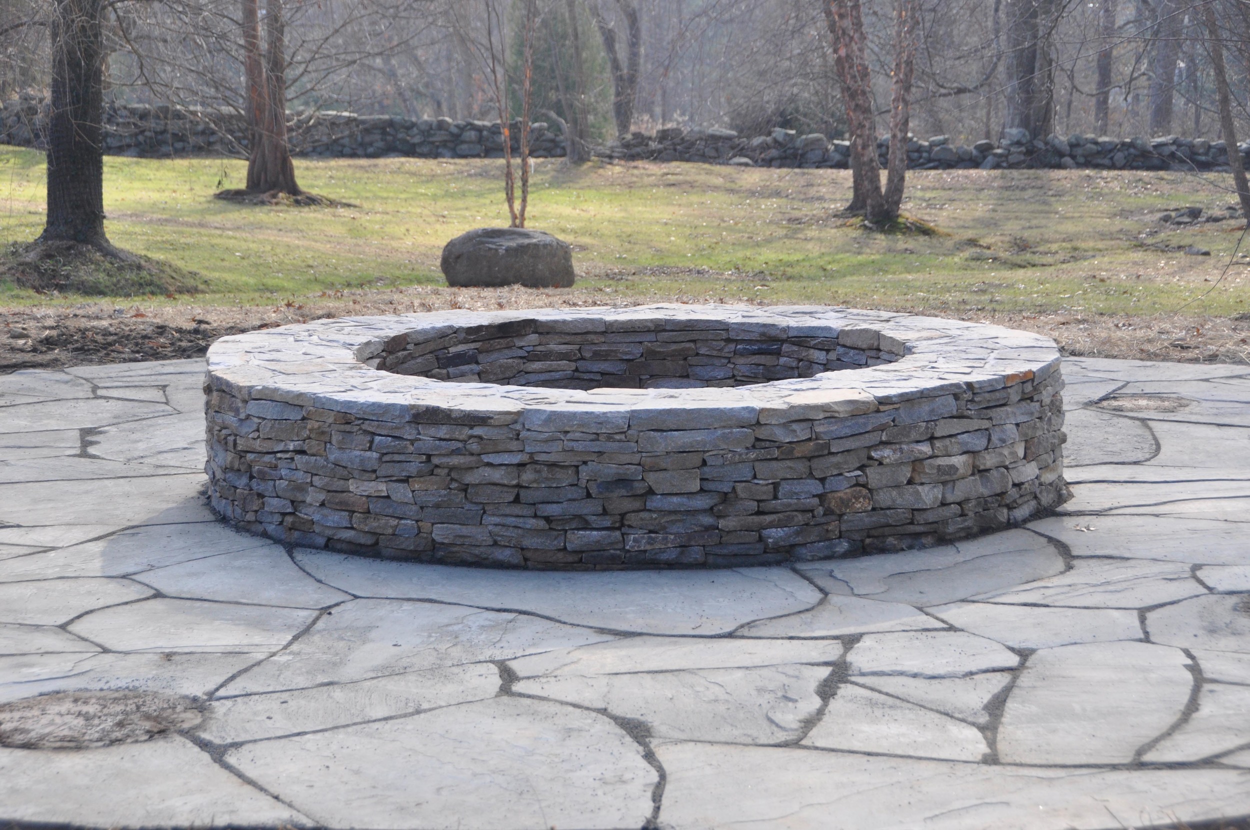 A circular stone fire pit with a layered stone wall, situated on a paved stone patio in a backyard with trees and grass in the background.