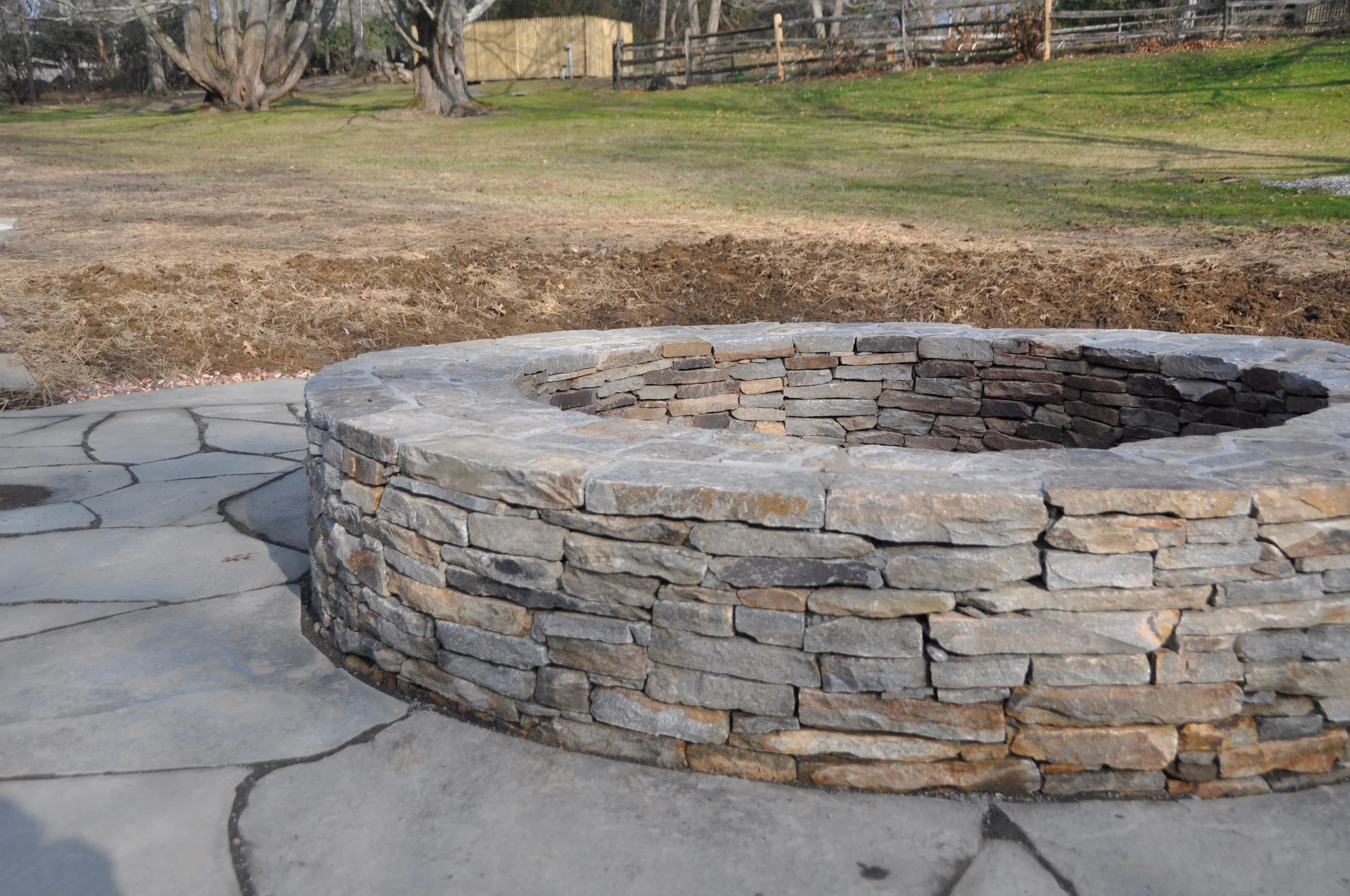 Stone fire pit on a concrete patio in a backyard with grass and trees in the background.
