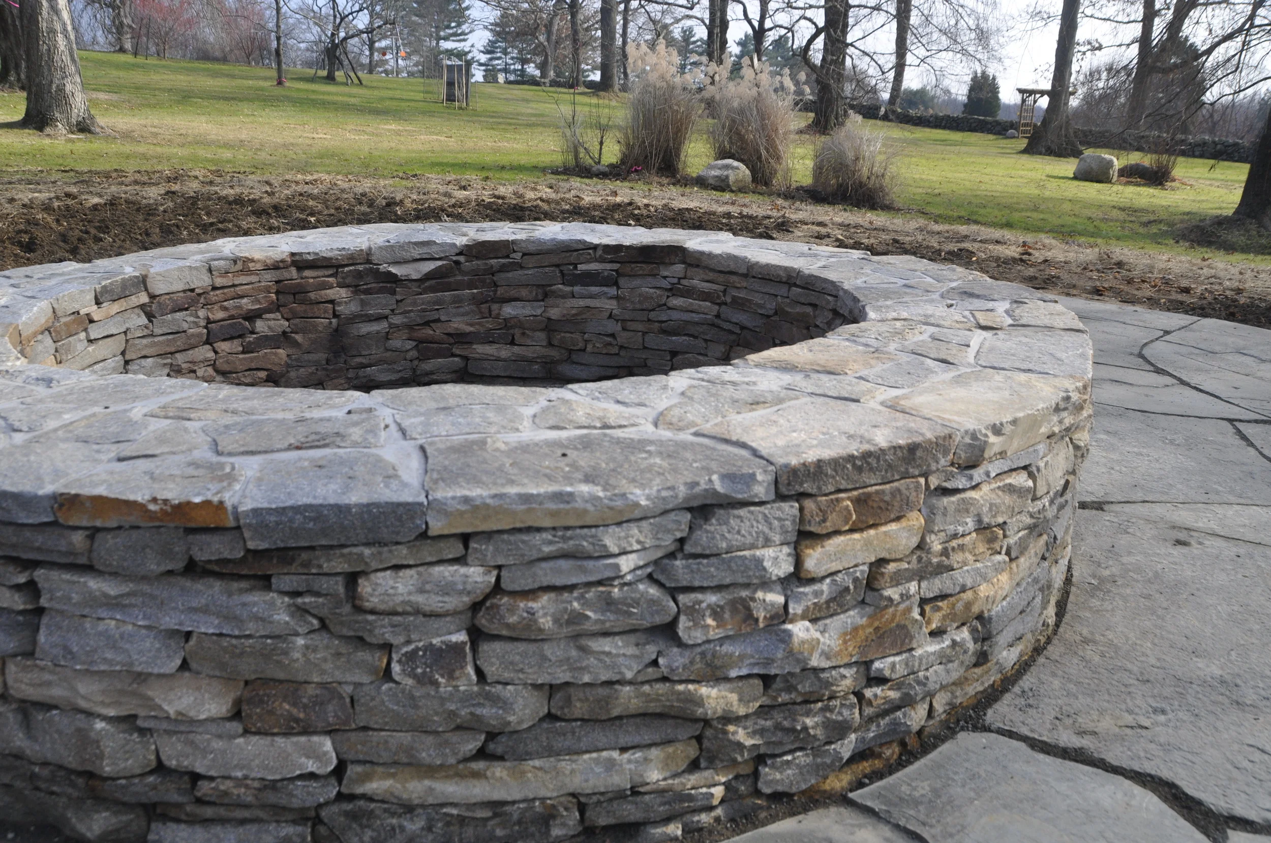 Stone fire pit surrounded by a concrete patio in a backyard with trees and grass.