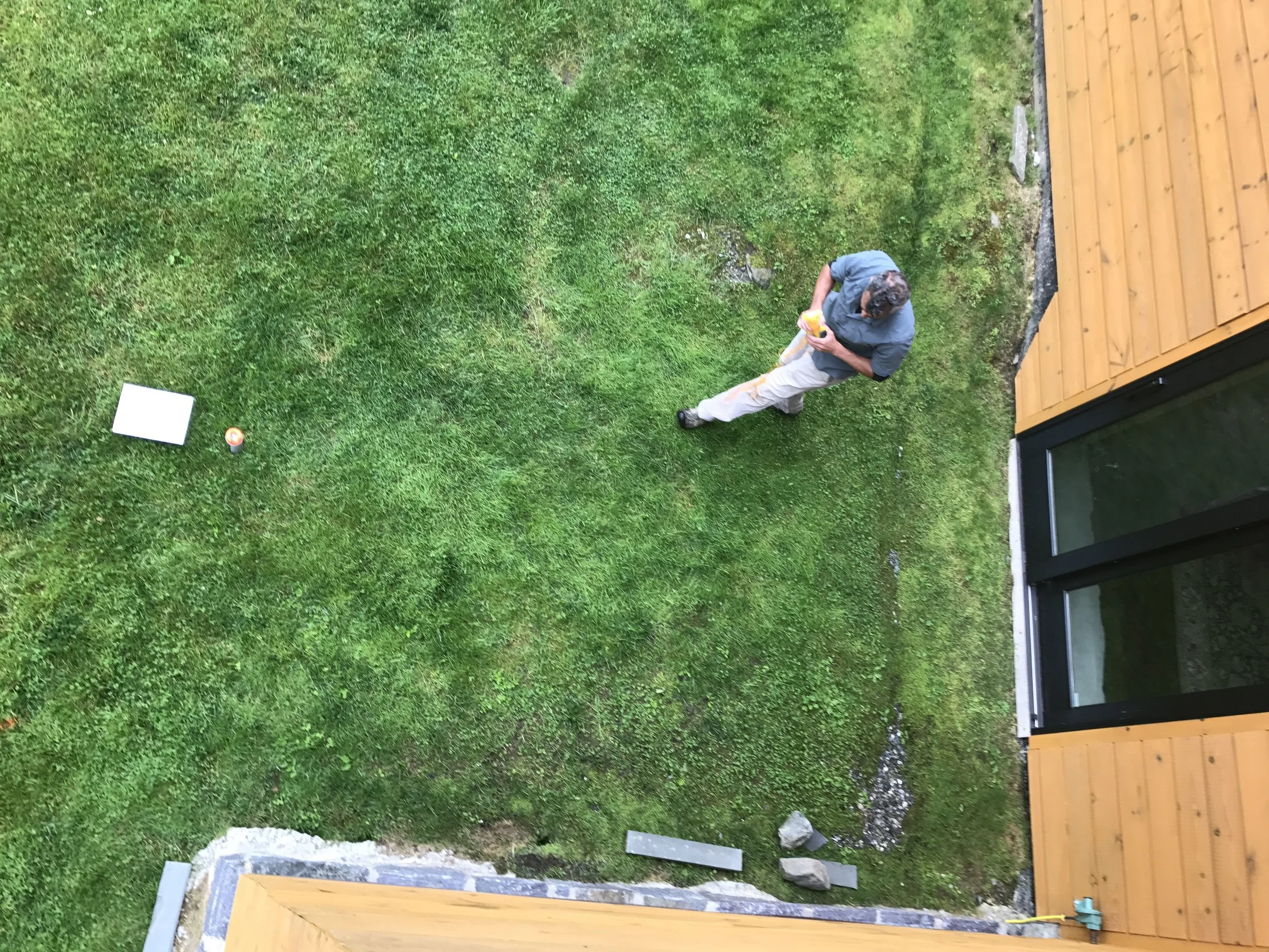 A man standing on grass next to a wooden building, holding a yellow and orange object, with construction materials nearby.