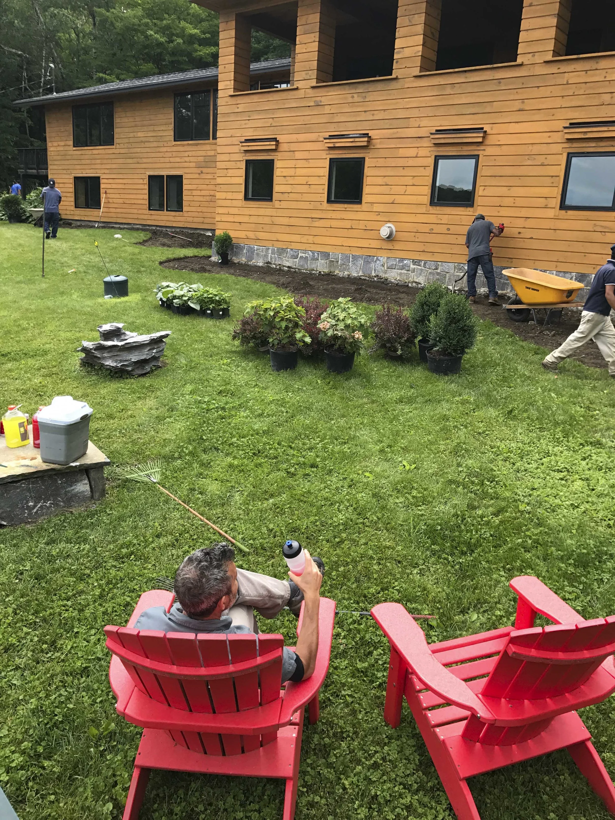 Man sitting on a red lawn chair in a yard, holding a drink, with two other red chairs nearby. In the background, workers are installing or finishing the exterior of a wooden house, with some tools and plants around.