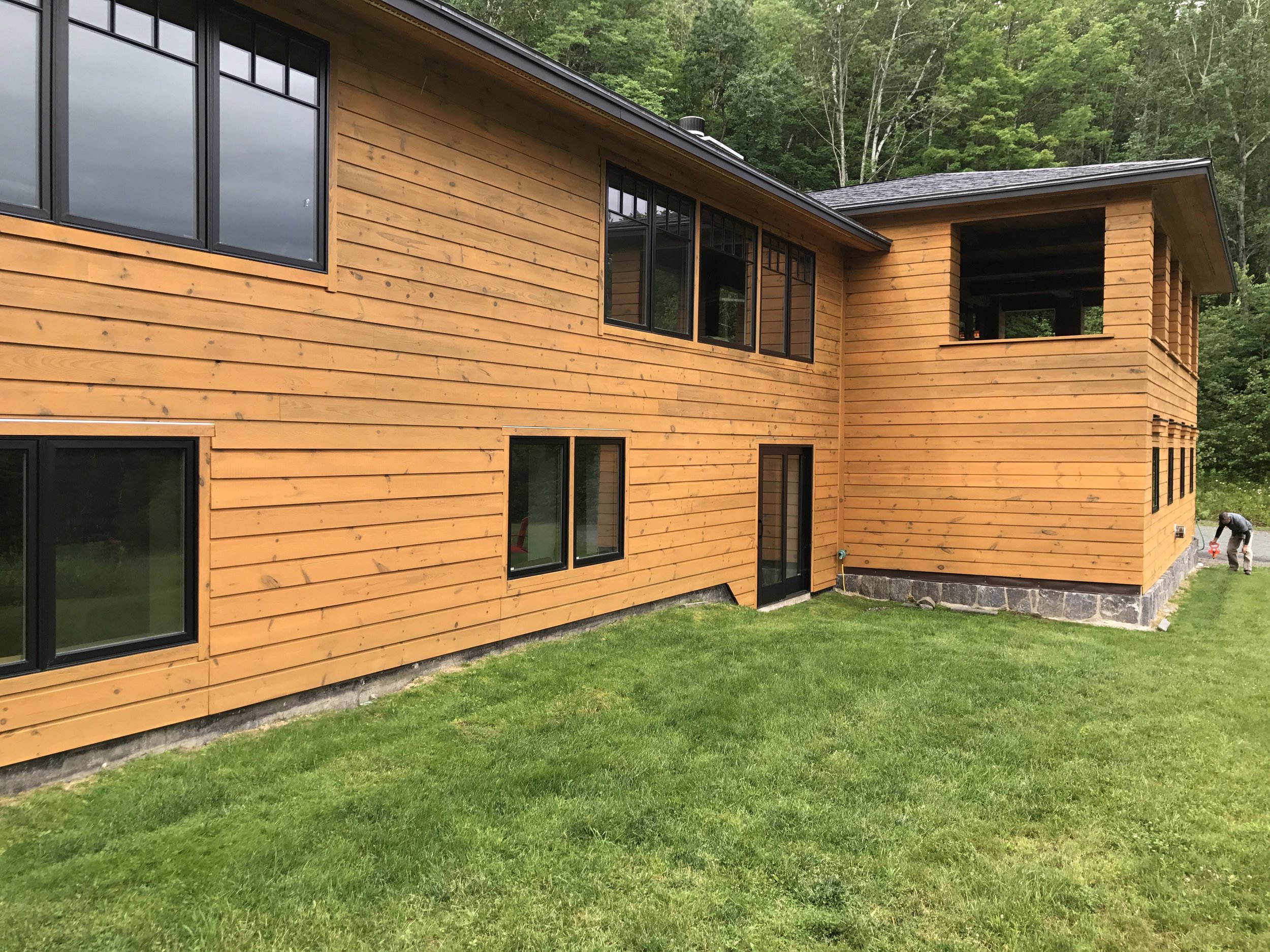 Newly constructed wooden house with large windows, situated in a grassy area with trees in the background. A person is working in the yard near the house on the right side.