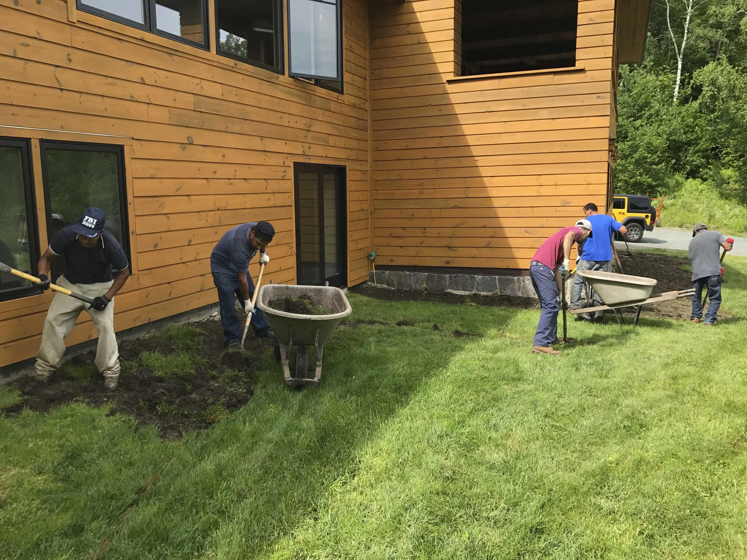 Group of workers planting grass and gardening around a wooden house with green grass and trees in the background.