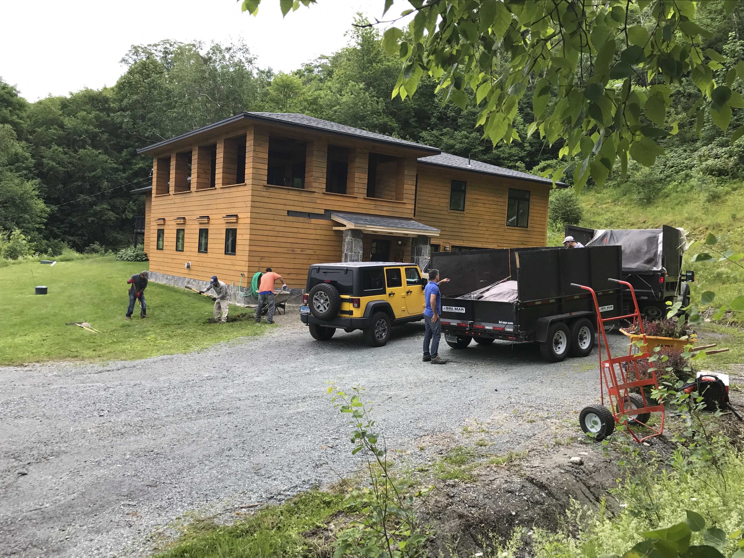 Construction workers are working on the yard of a two-story wooden house surrounded by greenery. A yellow Jeep and a black trailer are parked nearby, with equipment and tools around.