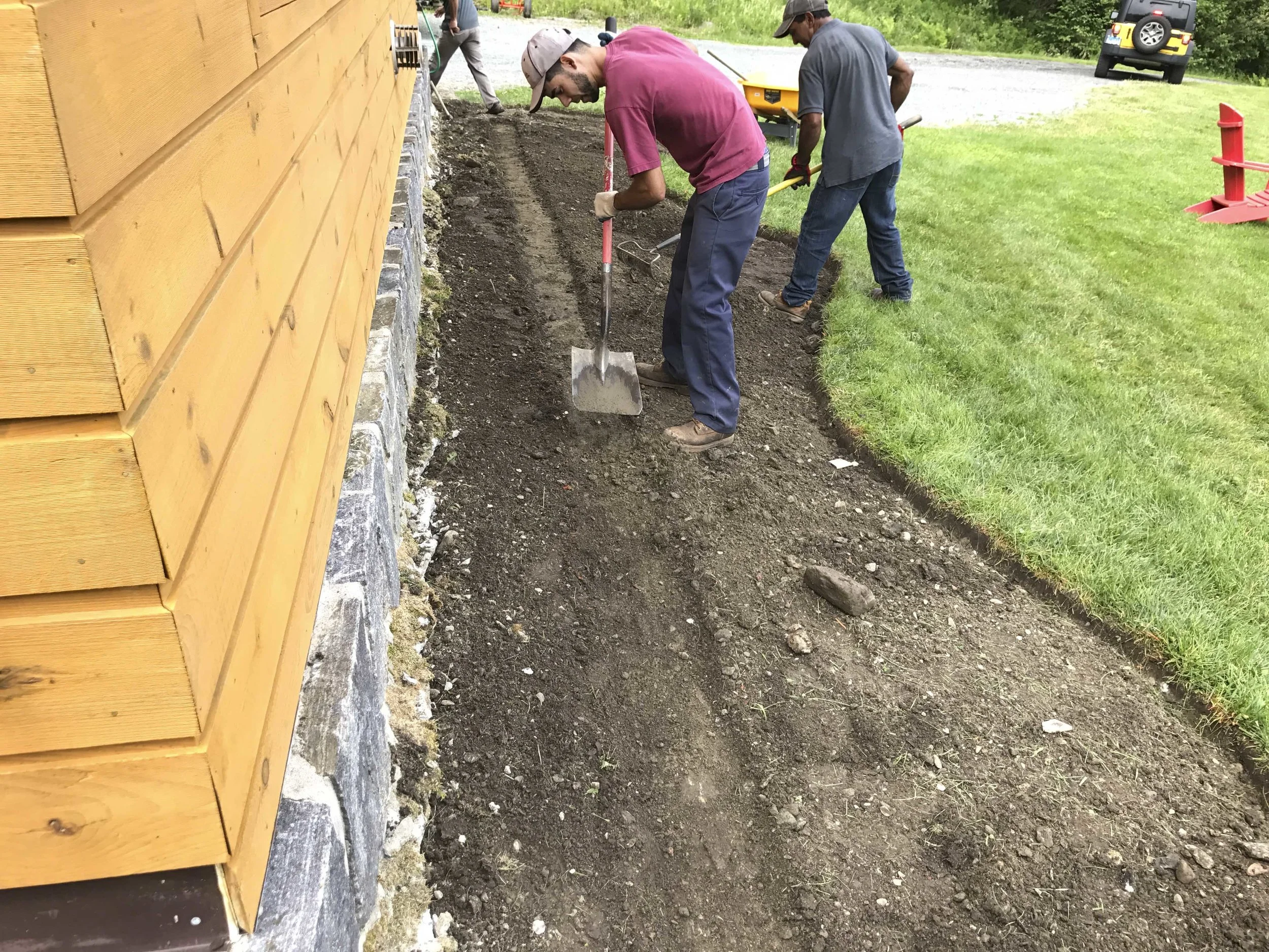 Three men working outside, leveling the ground along the side of a wooden building, using hand tools like a rake and a shovel, with a grassy yard and a car in the background.