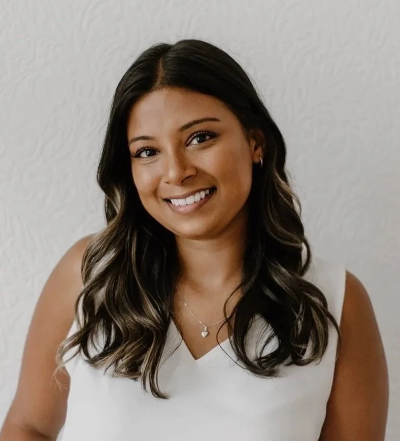 A young woman with long, dark wavy hair, smiling, wearing a white sleeveless top and a necklace with a heart-shaped pendant, standing against a light-colored textured wall.