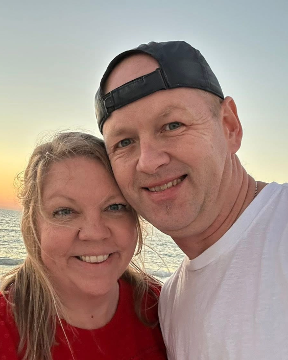 A smiling couple taking a selfie at the beach during sunset, with the ocean and sky in the background.
