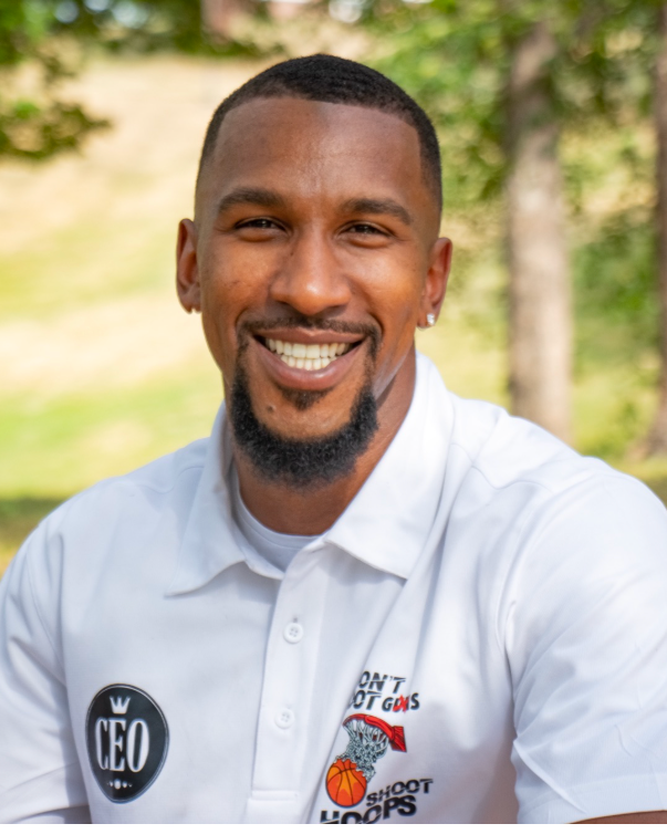 A smiling man with short hair and a goatee, wearing a white polo shirt with logos, outdoors with greenery in the background.