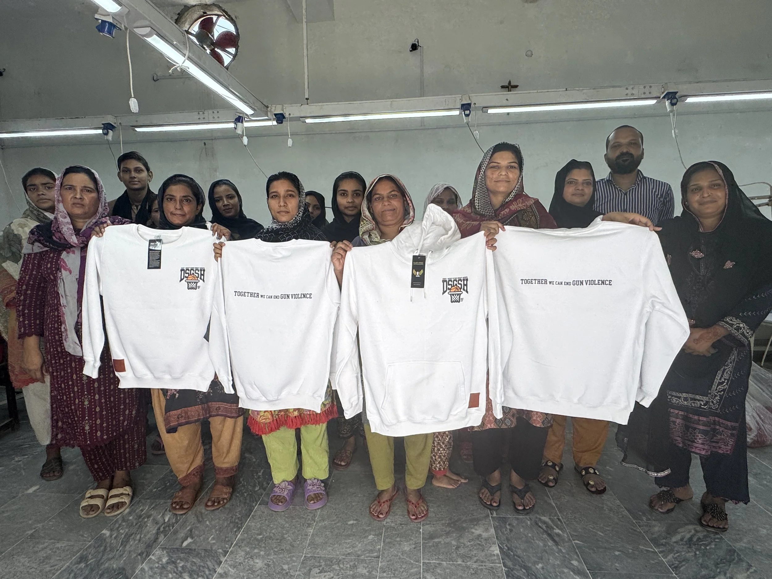Group of women and one man standing indoors, holding white T-shirts with a message that reads, 'TOGETHER WE CAN END GUN VIOLENCE.' The women are wearing traditional South Asian clothing and headscarves, and the man is wearing a striped shirt. The setting appears to be a community center or similar venue.