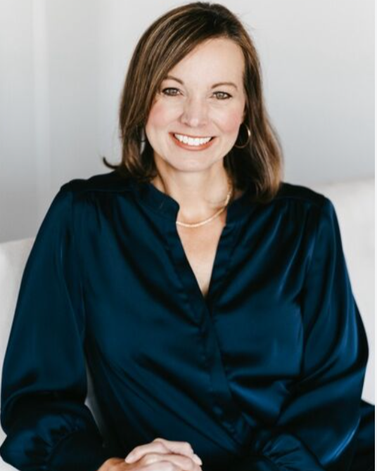 A woman with shoulder-length brown hair and a cheerful smile, wearing a dark blue satin blouse and gold jewelry, sitting against a plain light background.