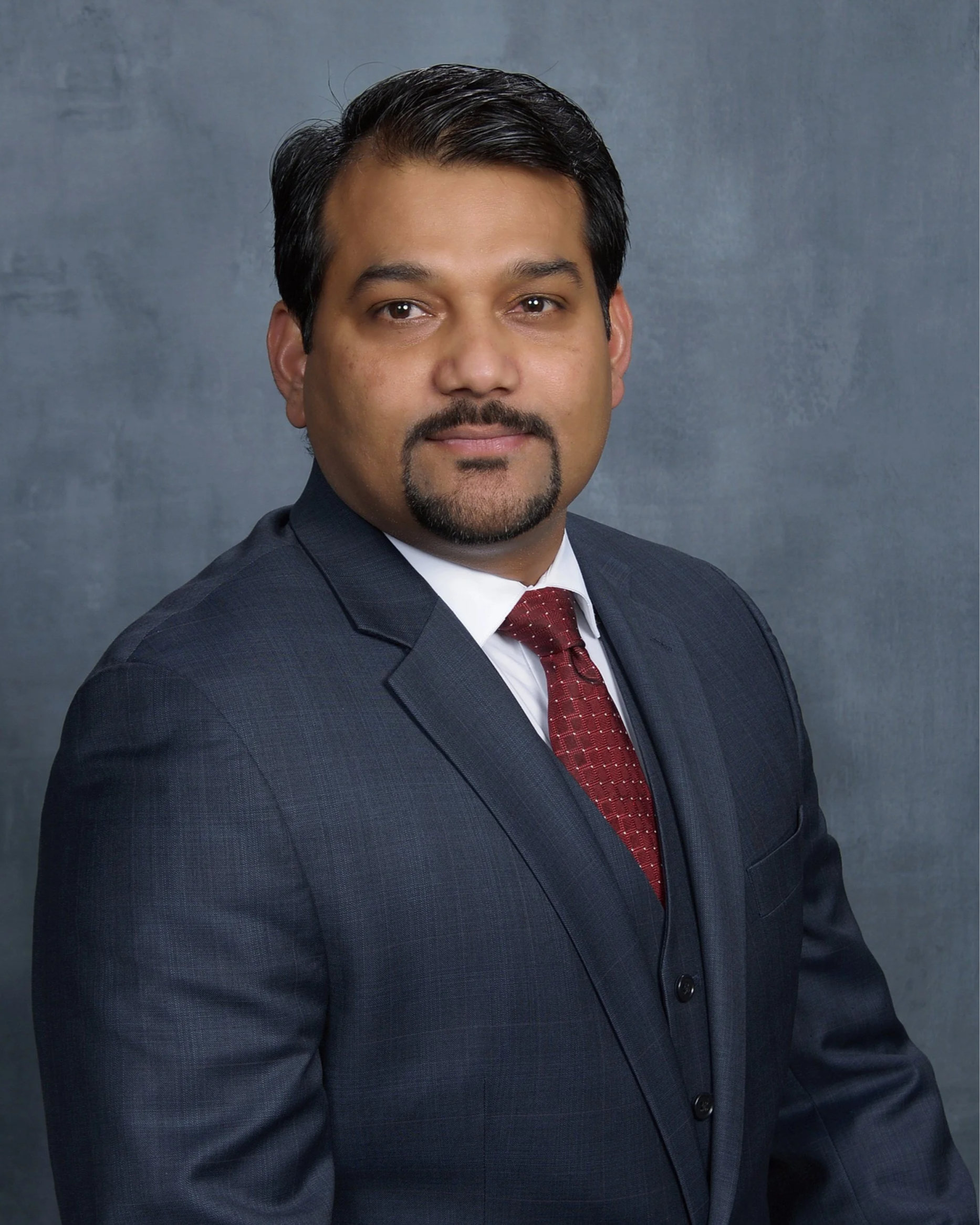 A man with dark hair, facial hair, wearing a dark suit, white shirt, and red tie, standing against a grey textured background.