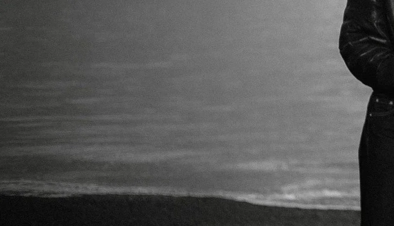 Close-up of a person wearing a black leather jacket standing on a beach at sunset, with the ocean waves in the background.