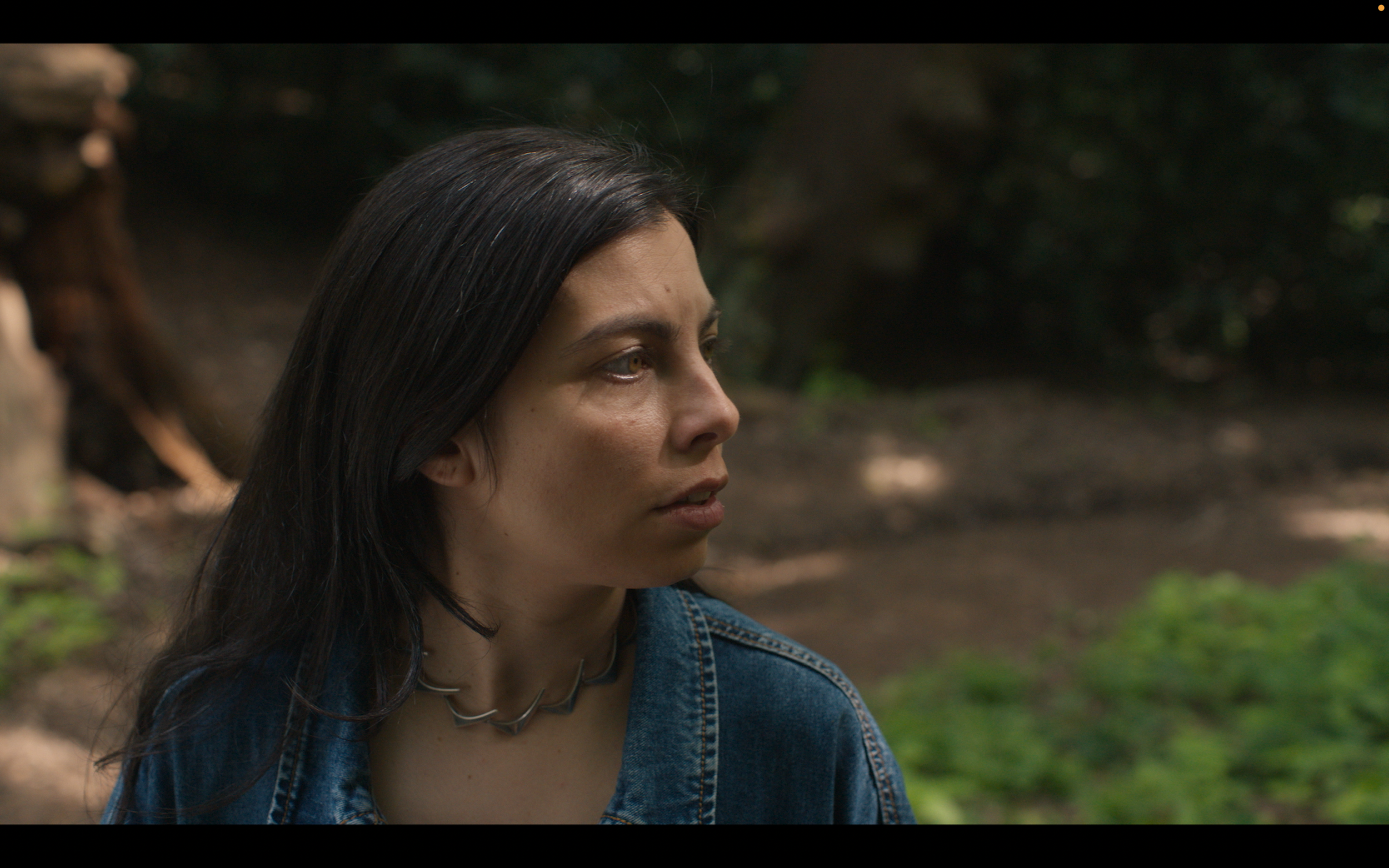 A woman with long dark hair, wearing a denim jacket and a necklace, is sitting outdoors in a forested area.