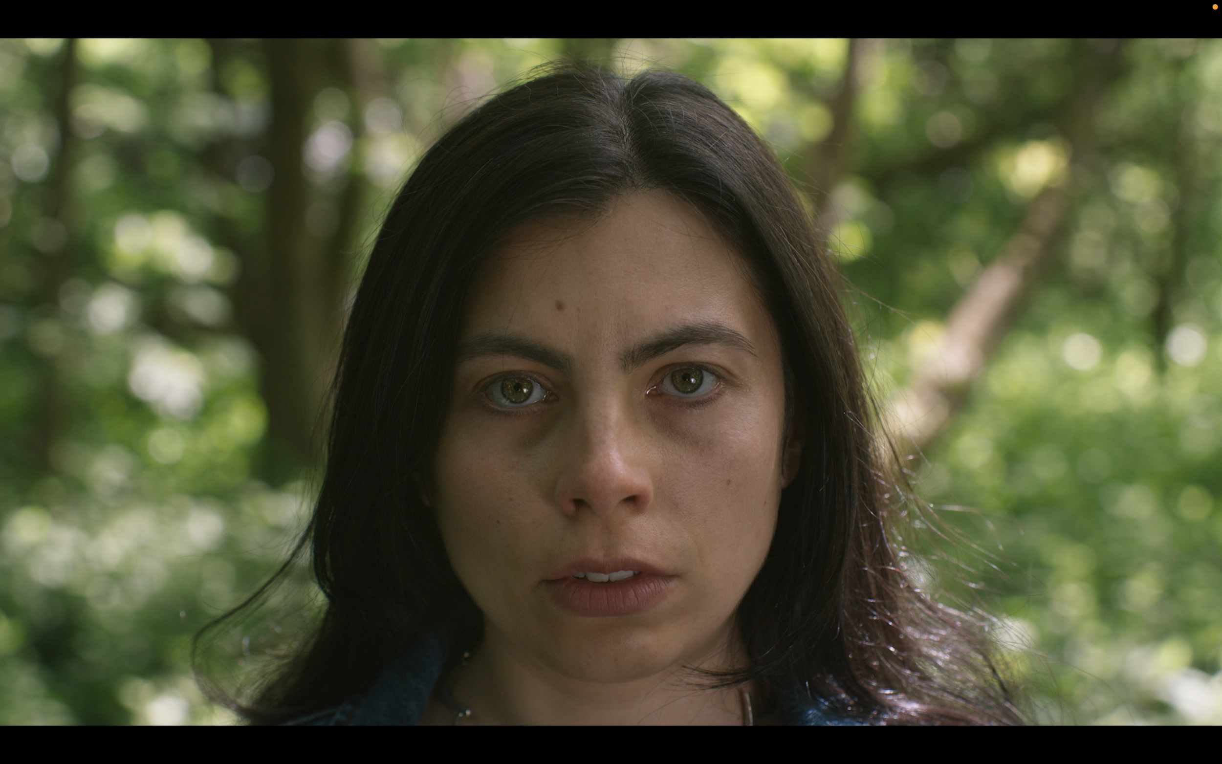 Close-up of a woman's face with dark hair and light-colored eyes, outdoors with blurred green foliage in the background.