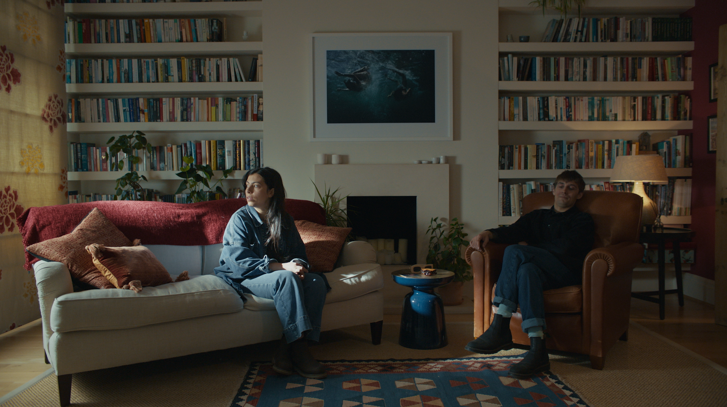 A woman and a man sitting in a living room with bookshelves, a framed picture of swimmers, and a lamp. The woman is on a beige sofa with pillows, and the man is in a brown armchair.