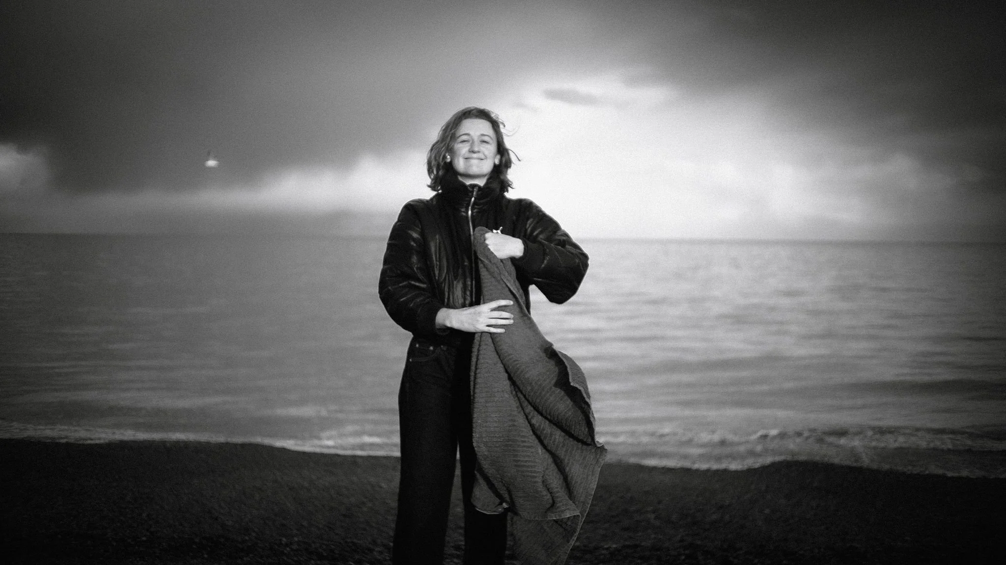 A woman smiling at the camera while standing on a beach with an ocean and cloudy sky in the background, in black and white.