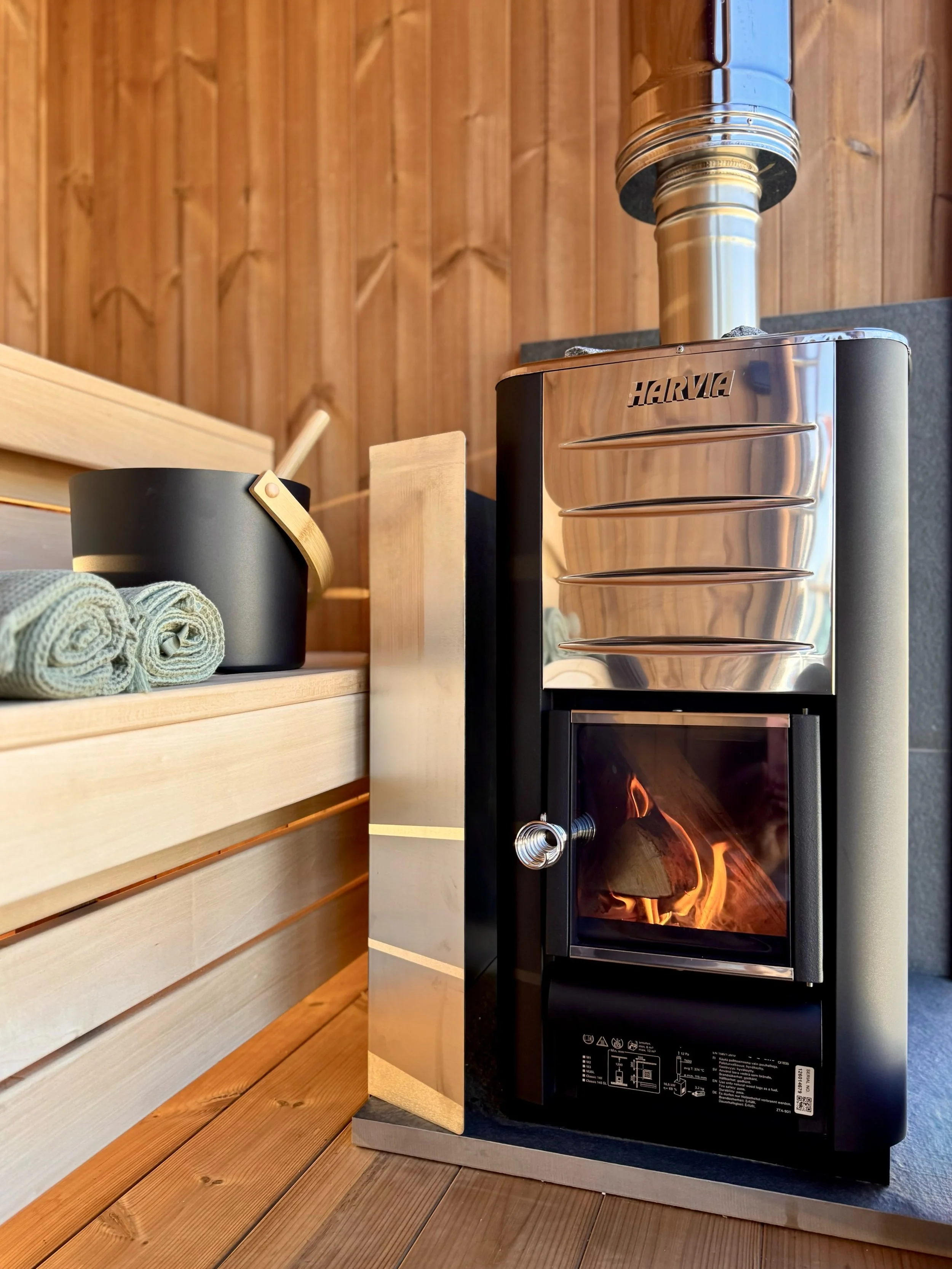 Inside of Still Wellness Sauna with Harvia stove lit showing flames, woodens auan benches with sauna bucket and sauna towels alongside, home sauna scotland