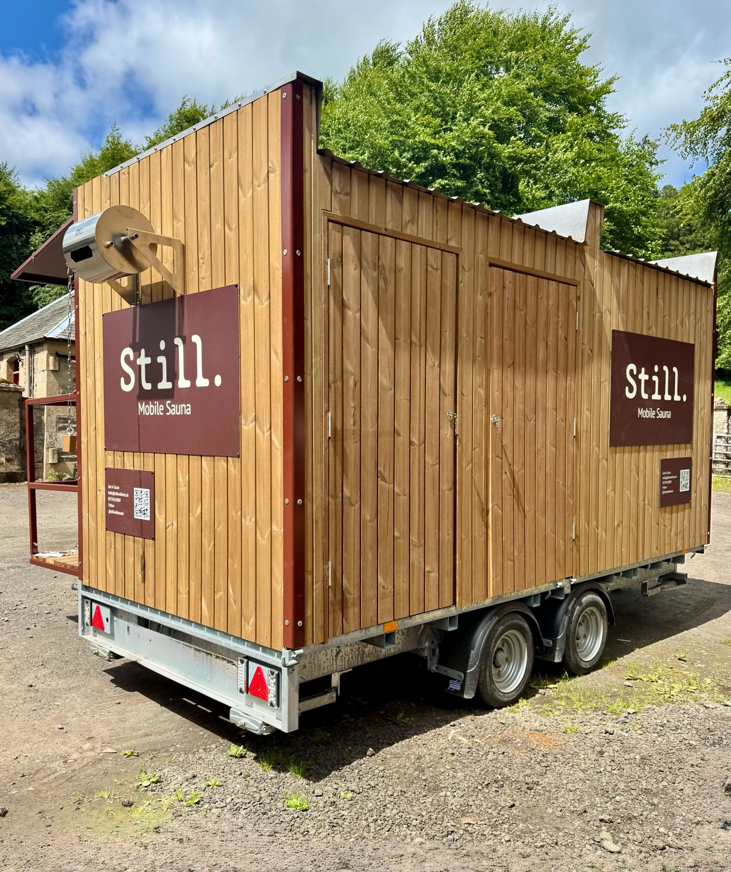 A mobile sauna on a trailer with wooden exterior walls and a roof, with signage reading "still. Mobile Sauna," parked on a gravel surface with green trees and a partly cloudy sky in the background.