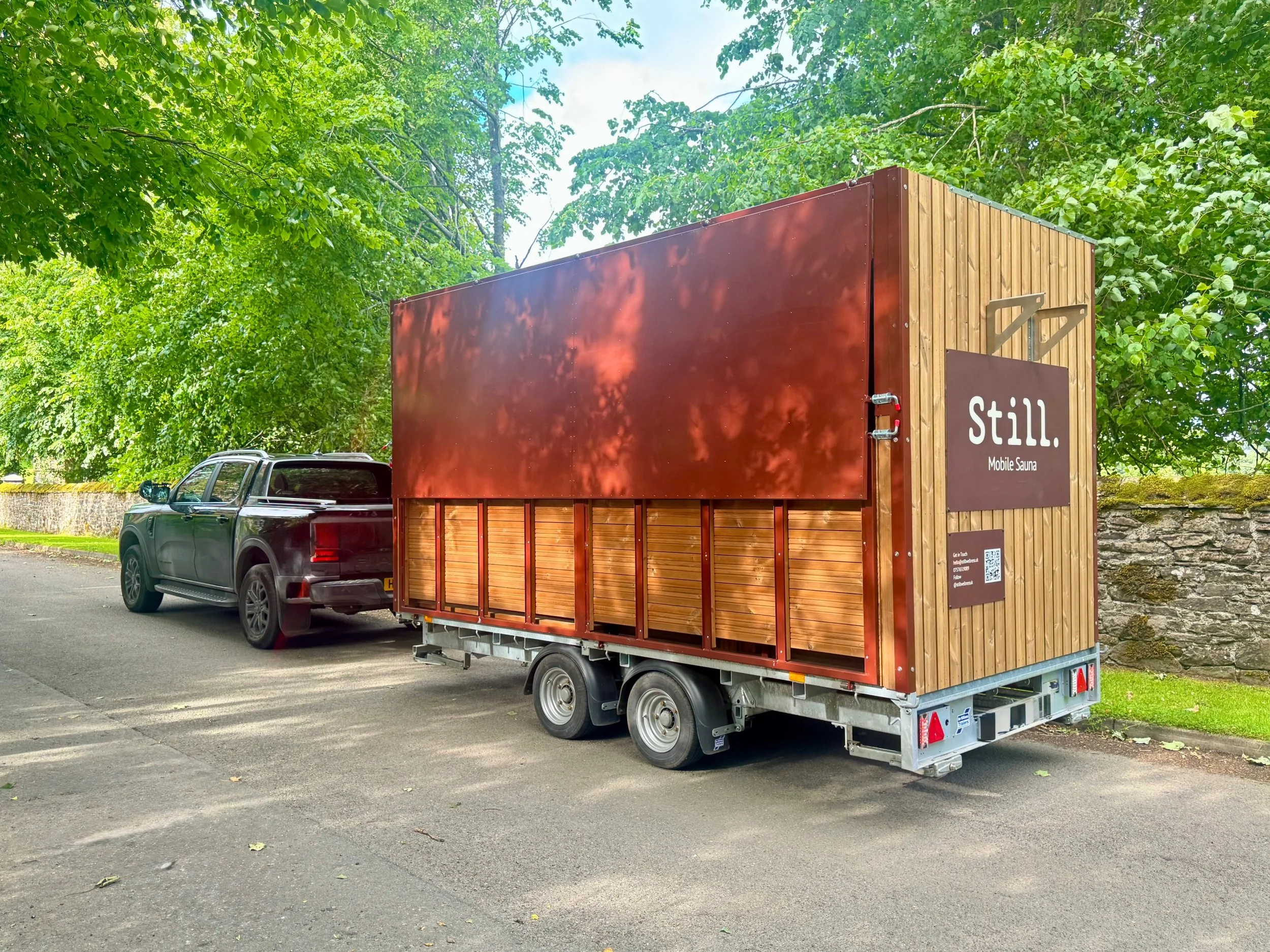 Mobile Sauna Hire Scotland by Still Wellness, Scotland, A fully mobile Sauna on a trailer pictured on tree framed road being driven by ford truck in Doune Scotland, Sauna Trailer design in brand colour Terracotta metalwork with thermawood cladding.