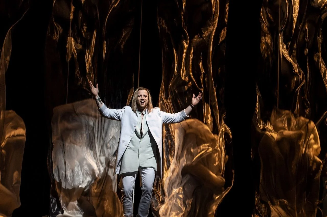 A man with long blonde hair, wearing a white suit, stands on stage with arms outstretched, smiling, in front of a dark background with swirling gold and black fabric.
