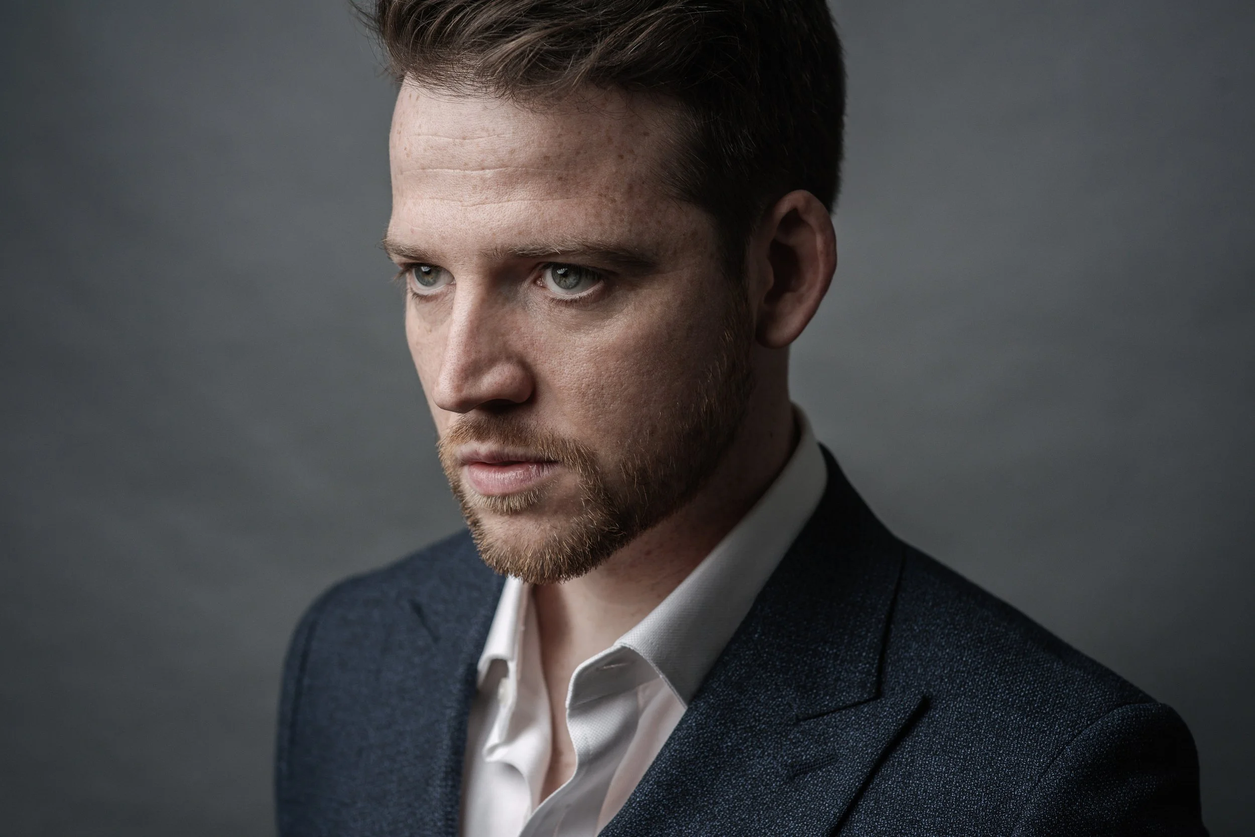 Close-up of a young man with blue eyes, light skin, brown hair, and a beard, wearing a dark blue suit jacket and a white shirt, looking contemplative against a gray background.