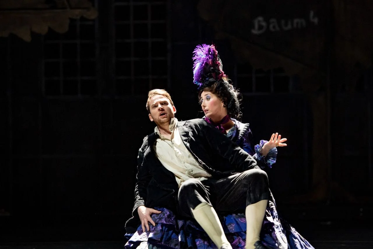 Two actors on stage performing in a theatrical play, with one man sitting and a woman in elaborate costume and feathered headpiece standing behind him.