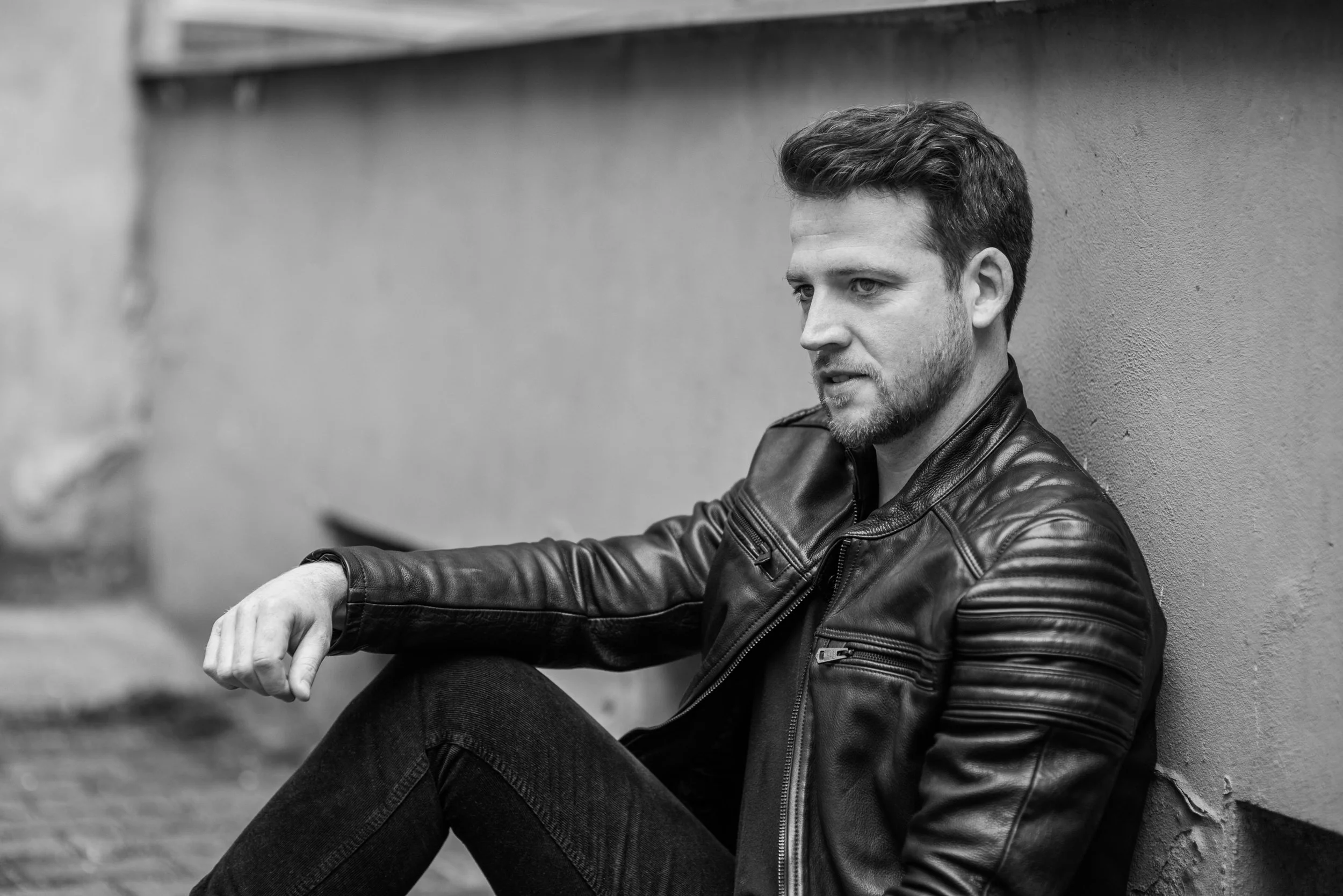 Black and white photo of a man with short hair and a beard, wearing a leather jacket, sitting against a wall with a thoughtful expression.