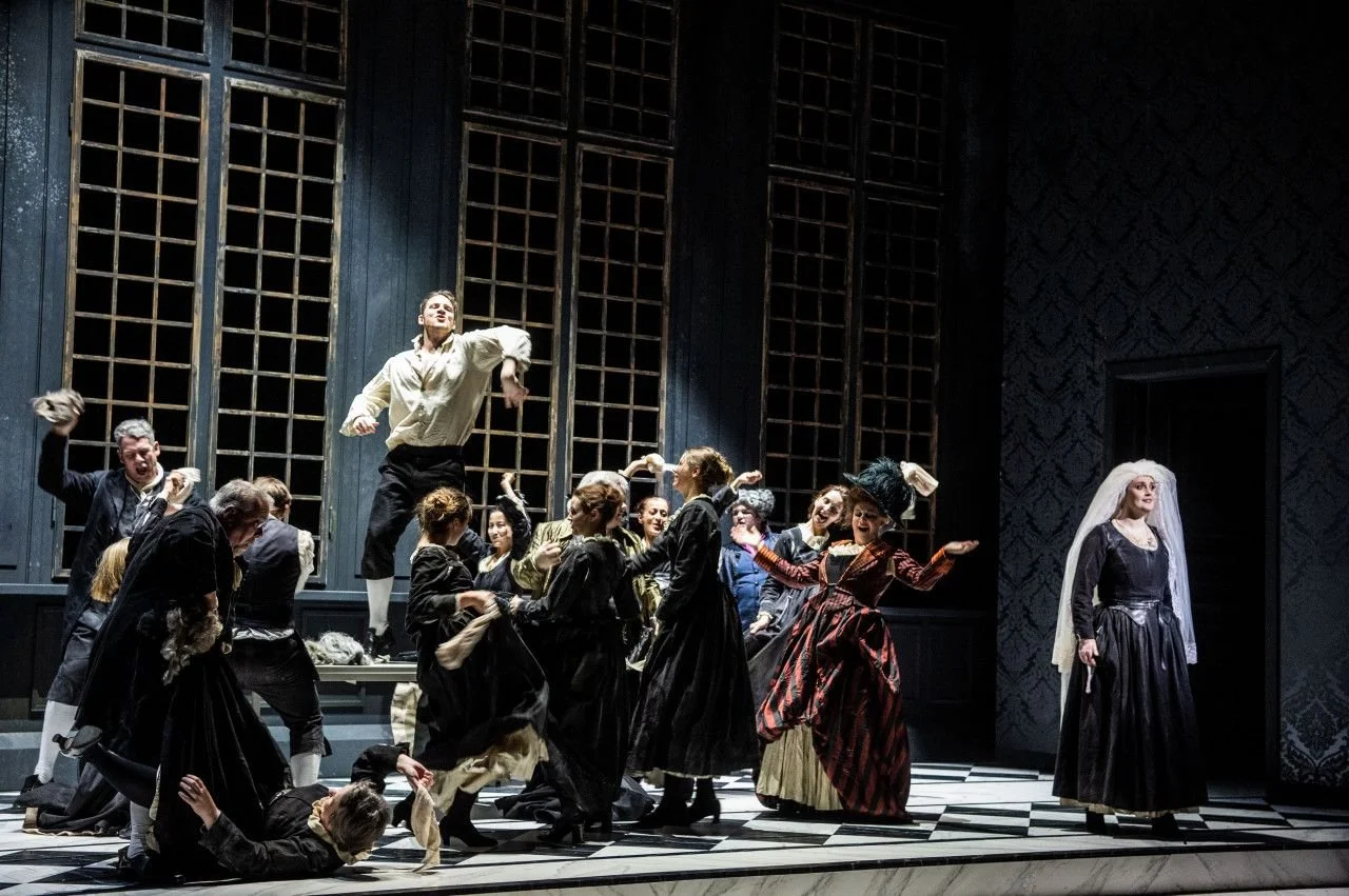 The image depicts a lively scene from a theatrical performance with actors dressed in period costumes, engaged in energetic dance or dialogue, on a stage with a patterned wall and large window-like structures in the background.