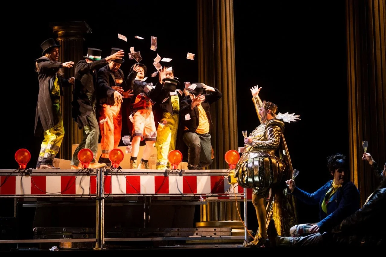 Stage performance with performers in colorful costumes, some in wizard hats, throwing money to a seated performer dressed in royal attire with a crown and golden outfit, surrounded by a black backdrop and stage decorations.