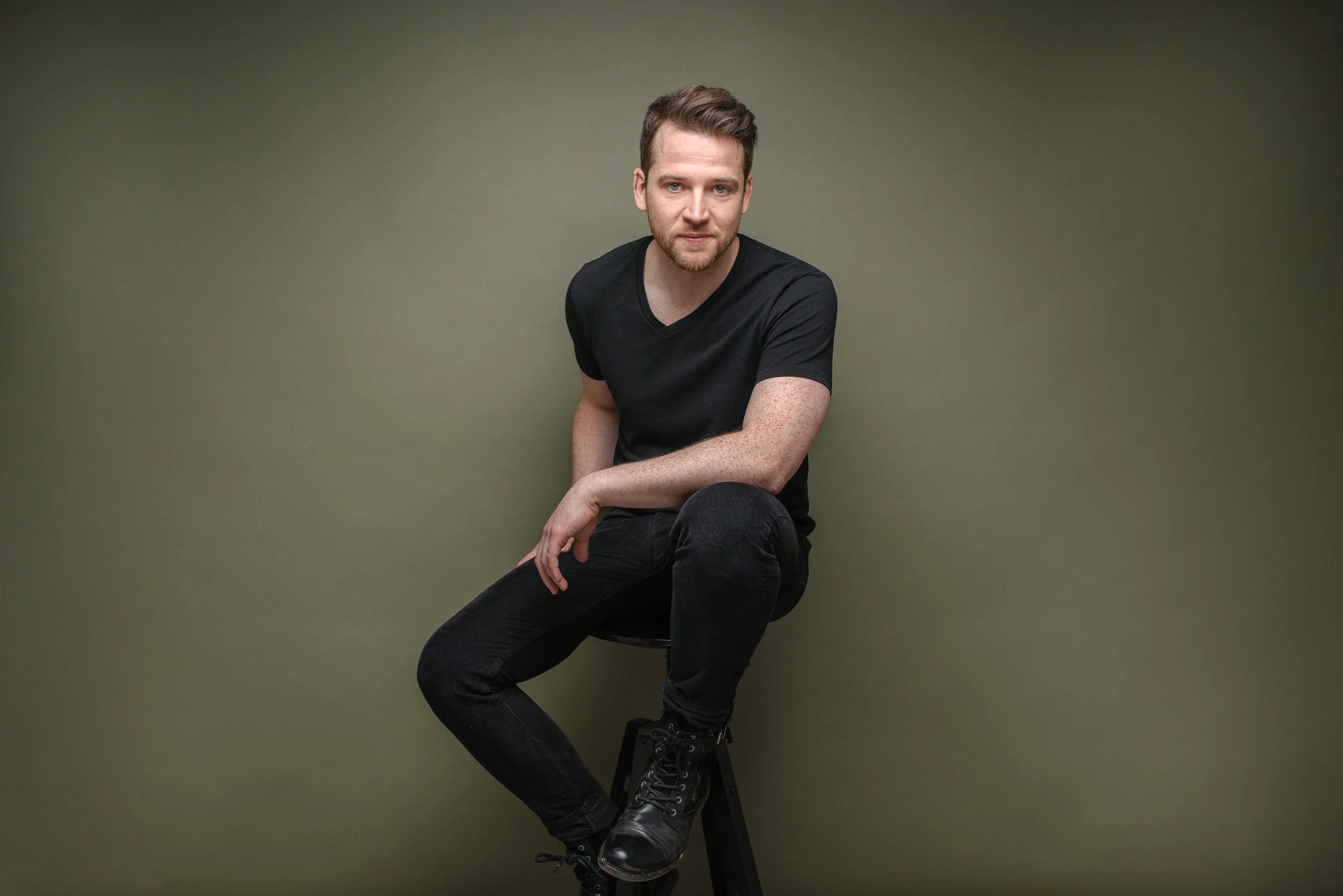 A man with short light brown hair and light skin, wearing a black t-shirt, black pants, and black boots, sitting on a stool against a plain olive green background.