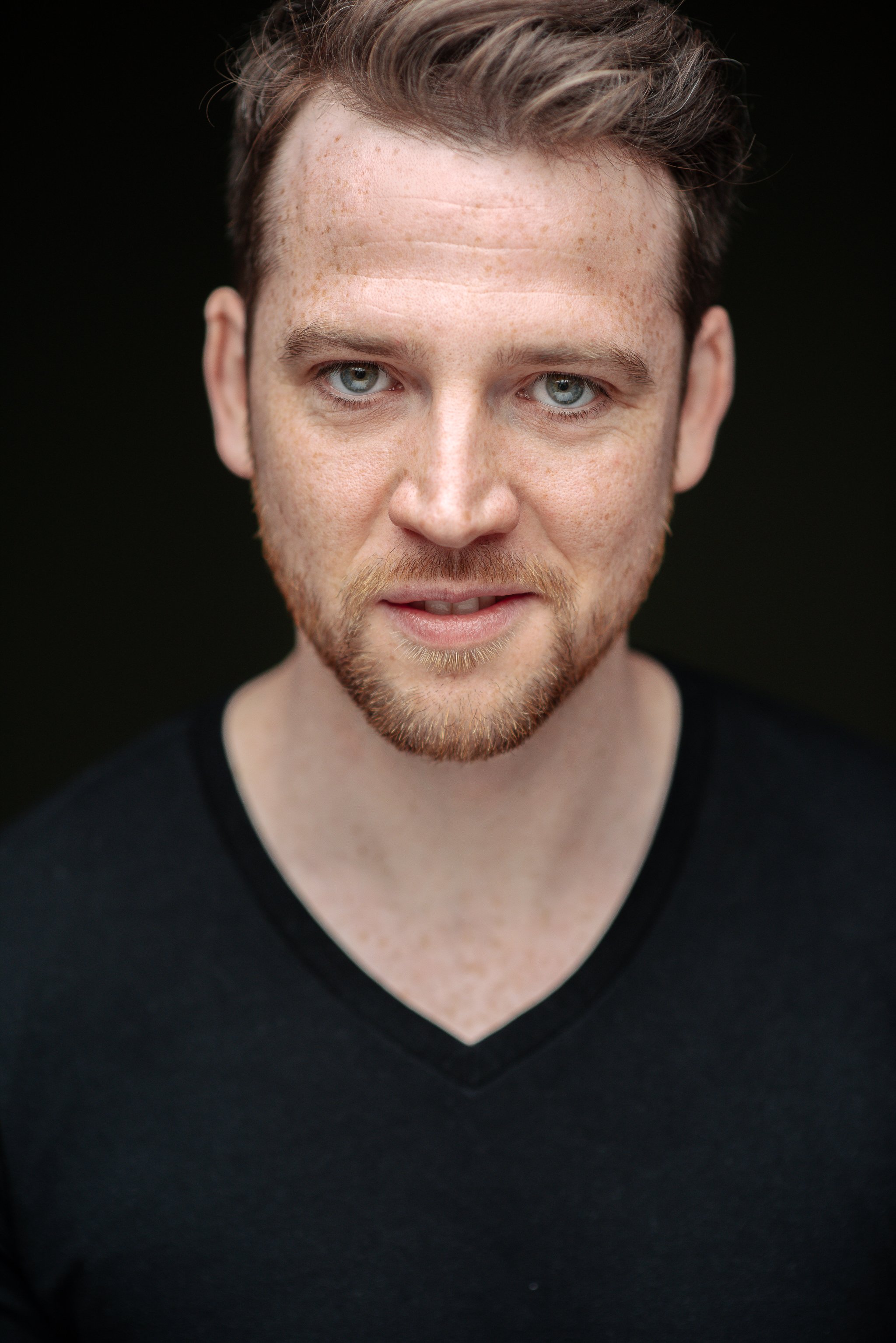 Close-up portrait of a young man with light skin, reddish hair, light blue eyes, and a short beard, wearing a black V-neck shirt, against a dark background.