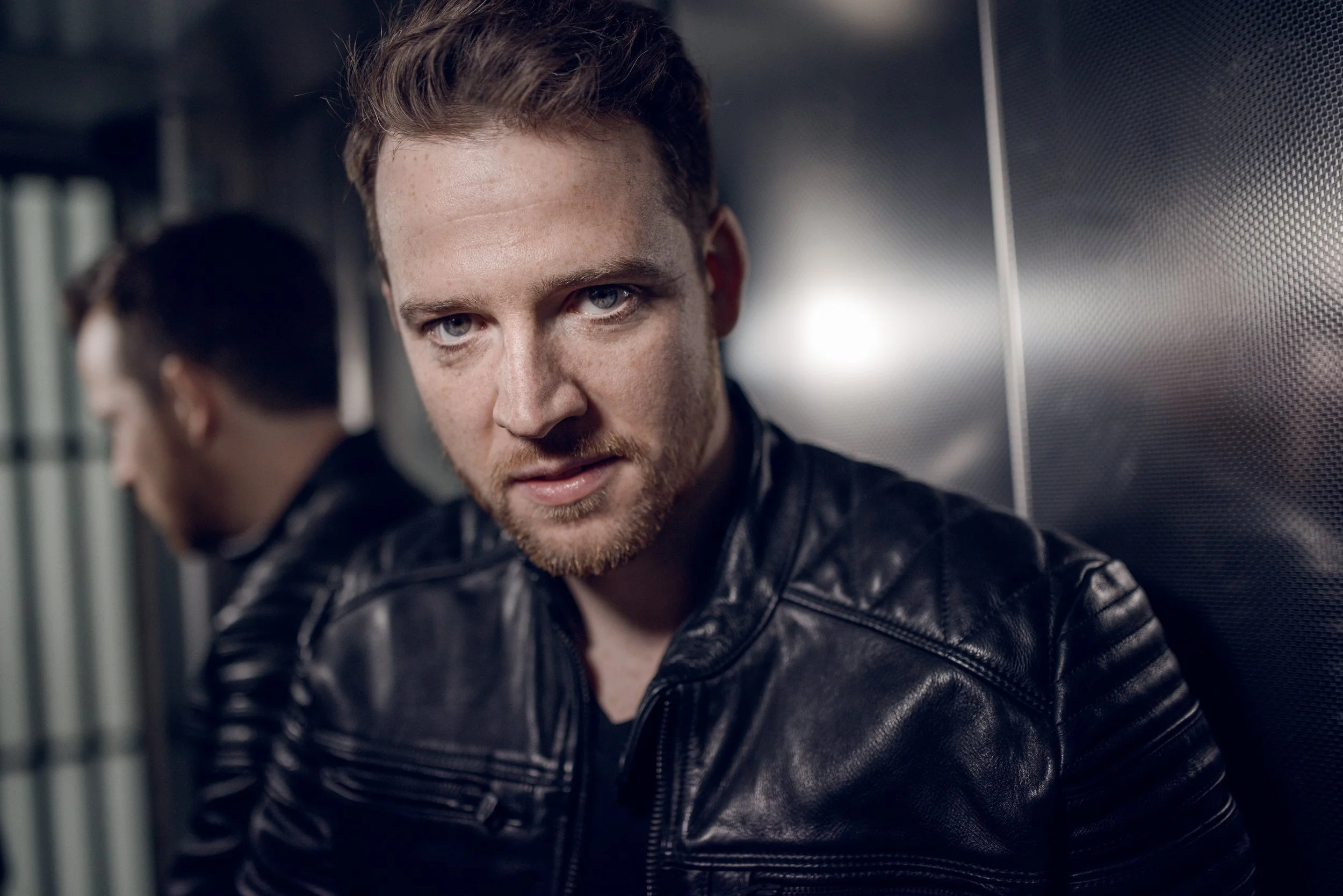 A young man with red hair and blue eyes wearing a black leather jacket, leaning against a reflective metal surface with his reflection visible.