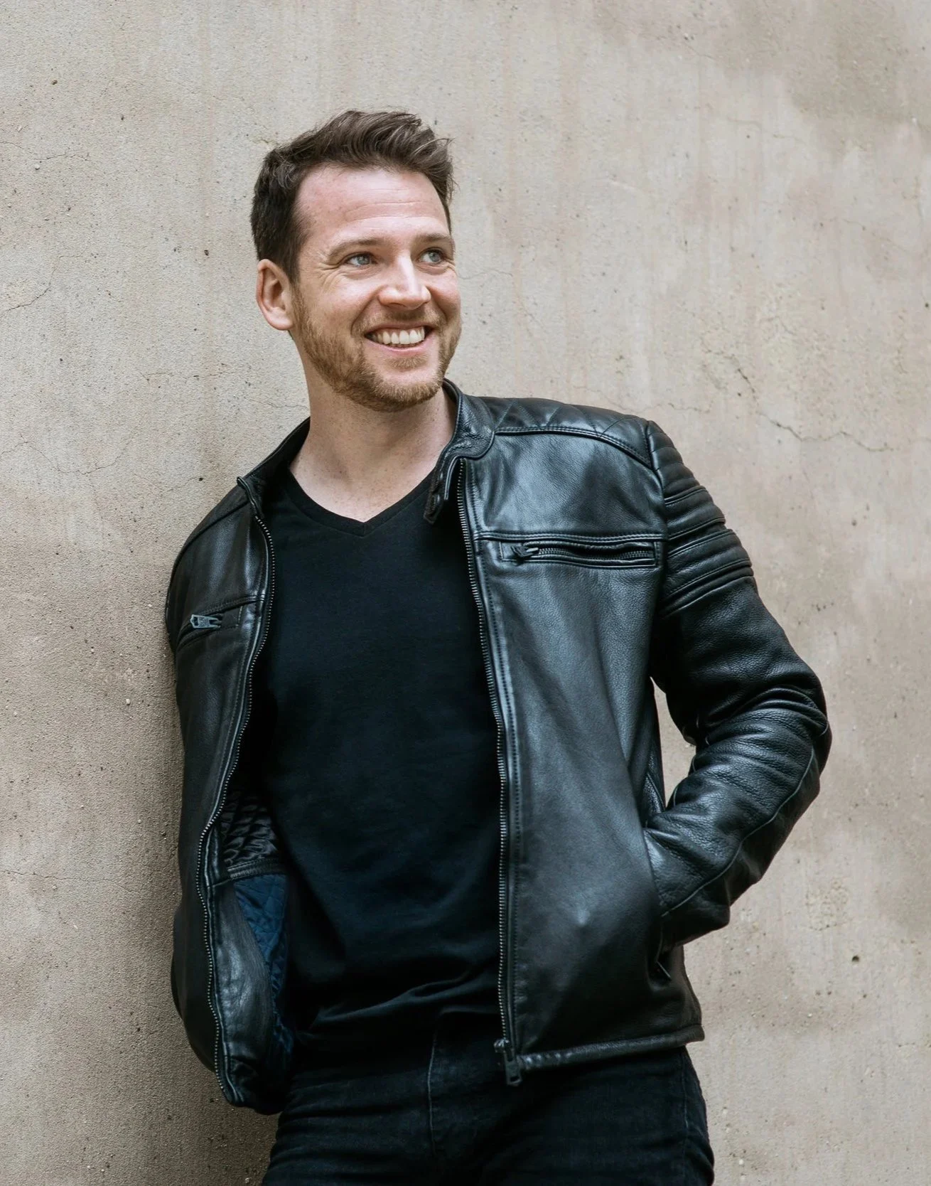 A man leaning against a beige wall, smiling, wearing a black leather jacket and black shirt.