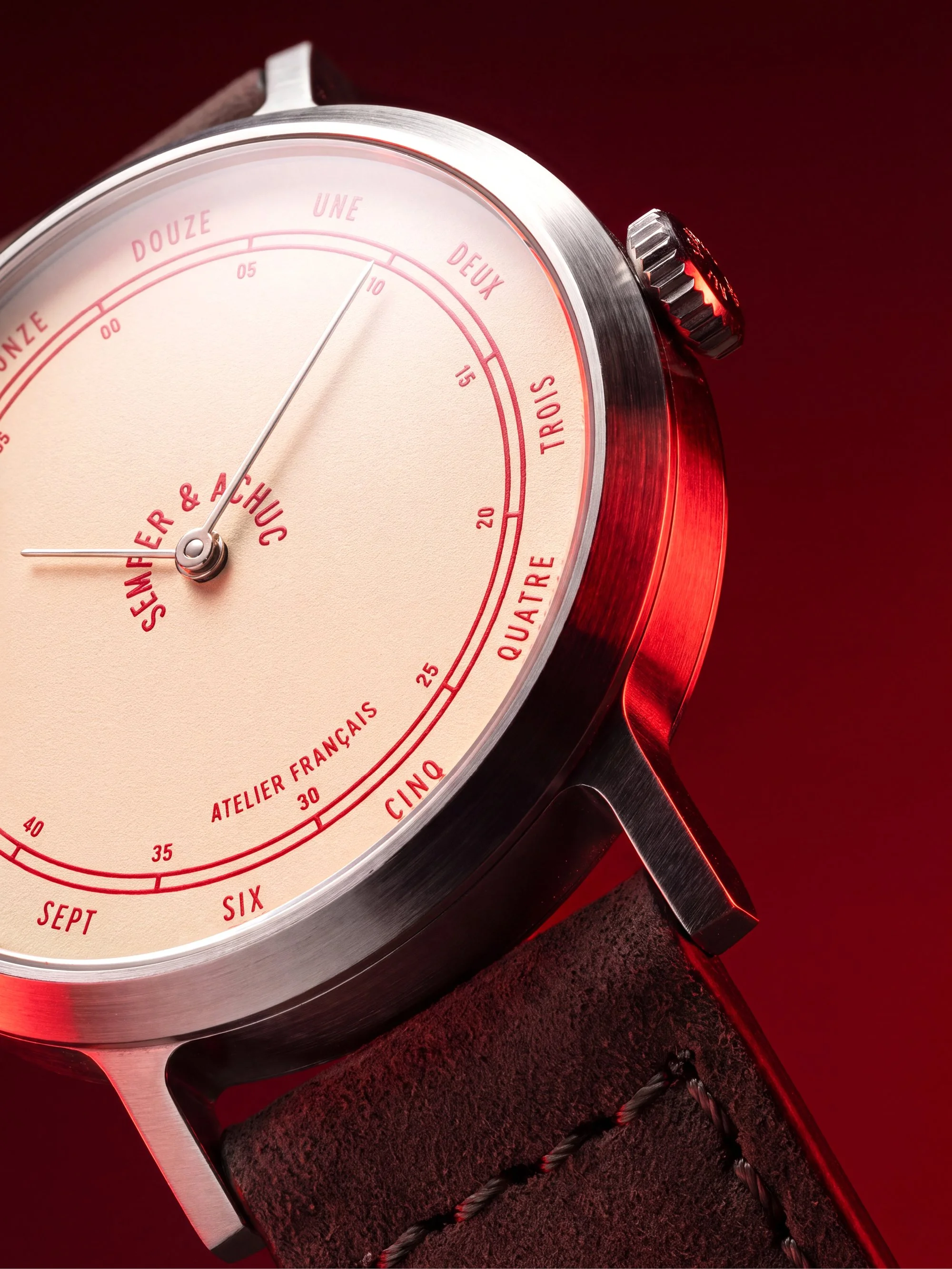 Close-up of a wristwatch with a beige dial, displaying months in French, with a silver bezel, black leather strap, and a textured surface, against a red background.
