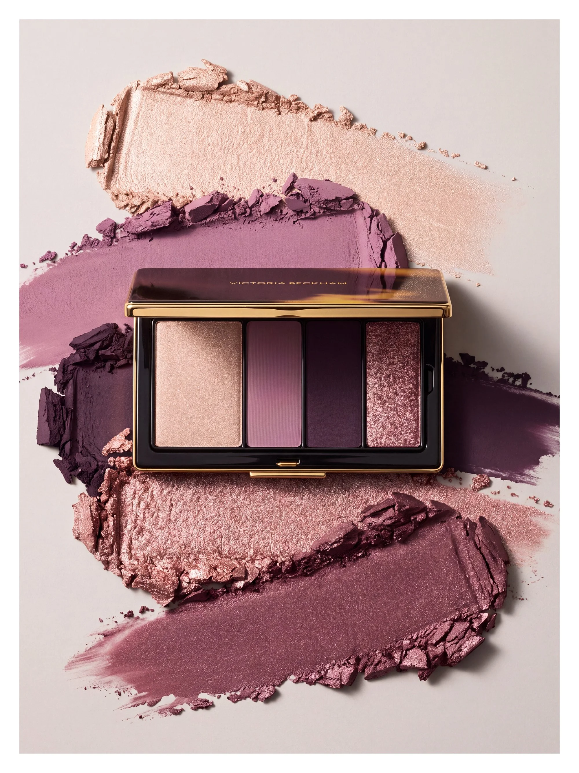 A makeup palette with four shades of pink and purple eyeshadow, placed on a background of crushed eyeshadows in similar shades, creating a colorful and artistic display.
