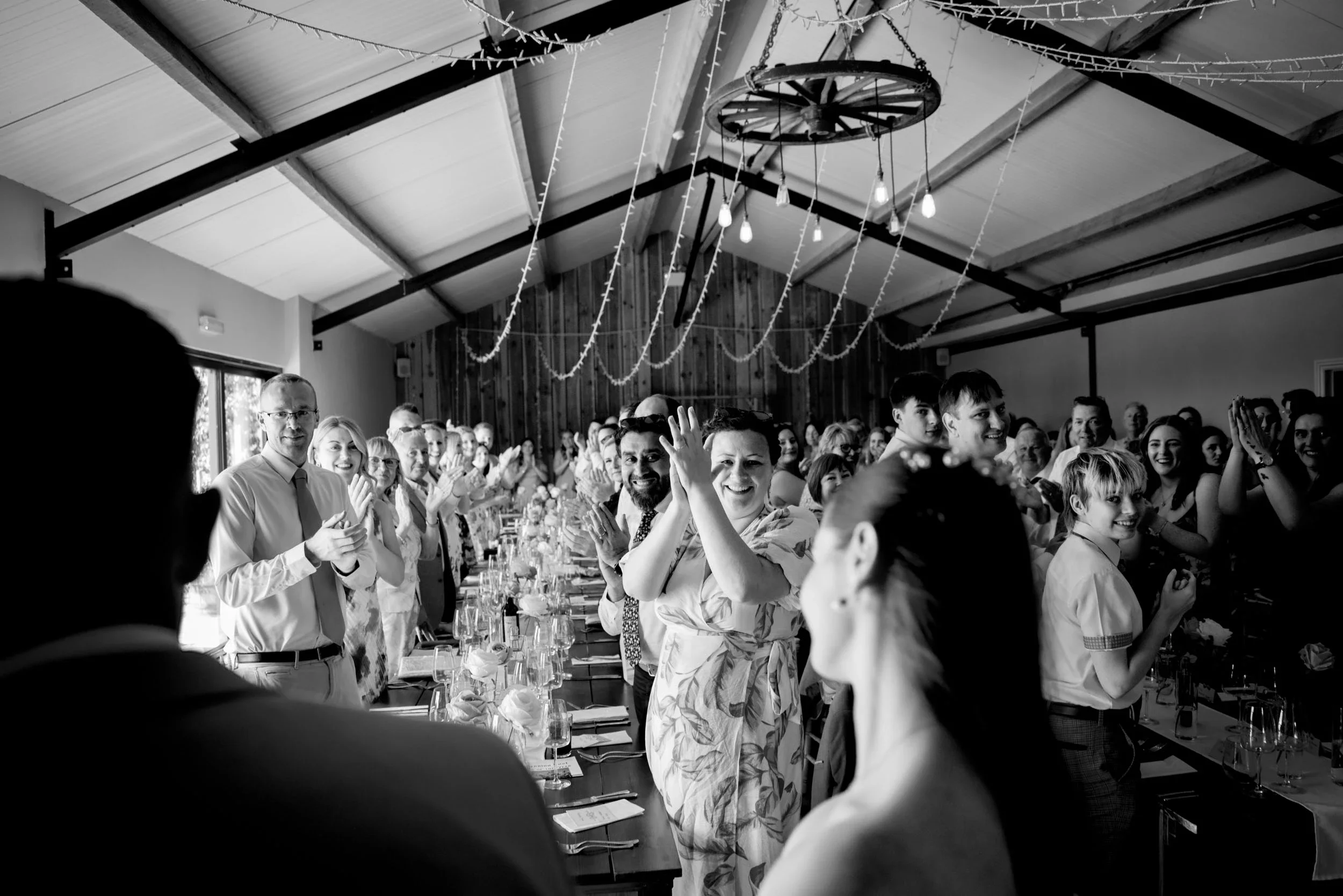 People standing around a long dining table at a celebration event, clapping and smiling, with decorations hanging from the ceiling in a large wooden hall.