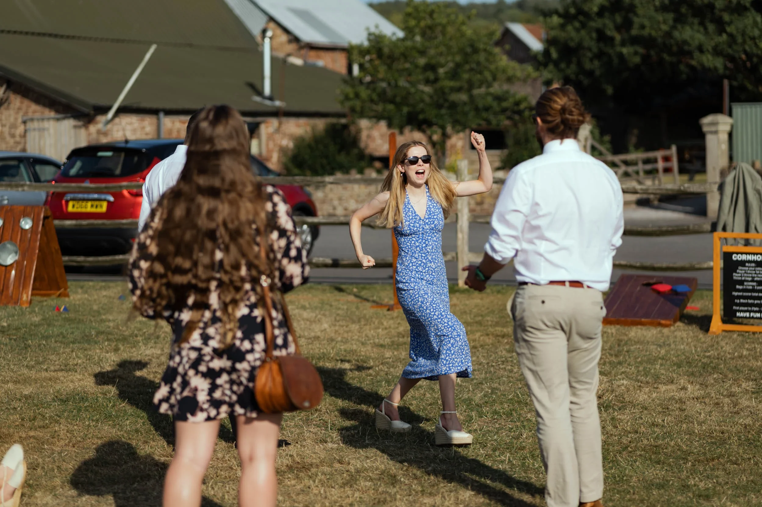 A woman in a blue floral dress and sunglasses is celebrating with a joyful expression and raised fist, while standing on a pair of high-heeled wedge shoes at an outdoor event. Other people are in the foreground and background, with cars and houses in