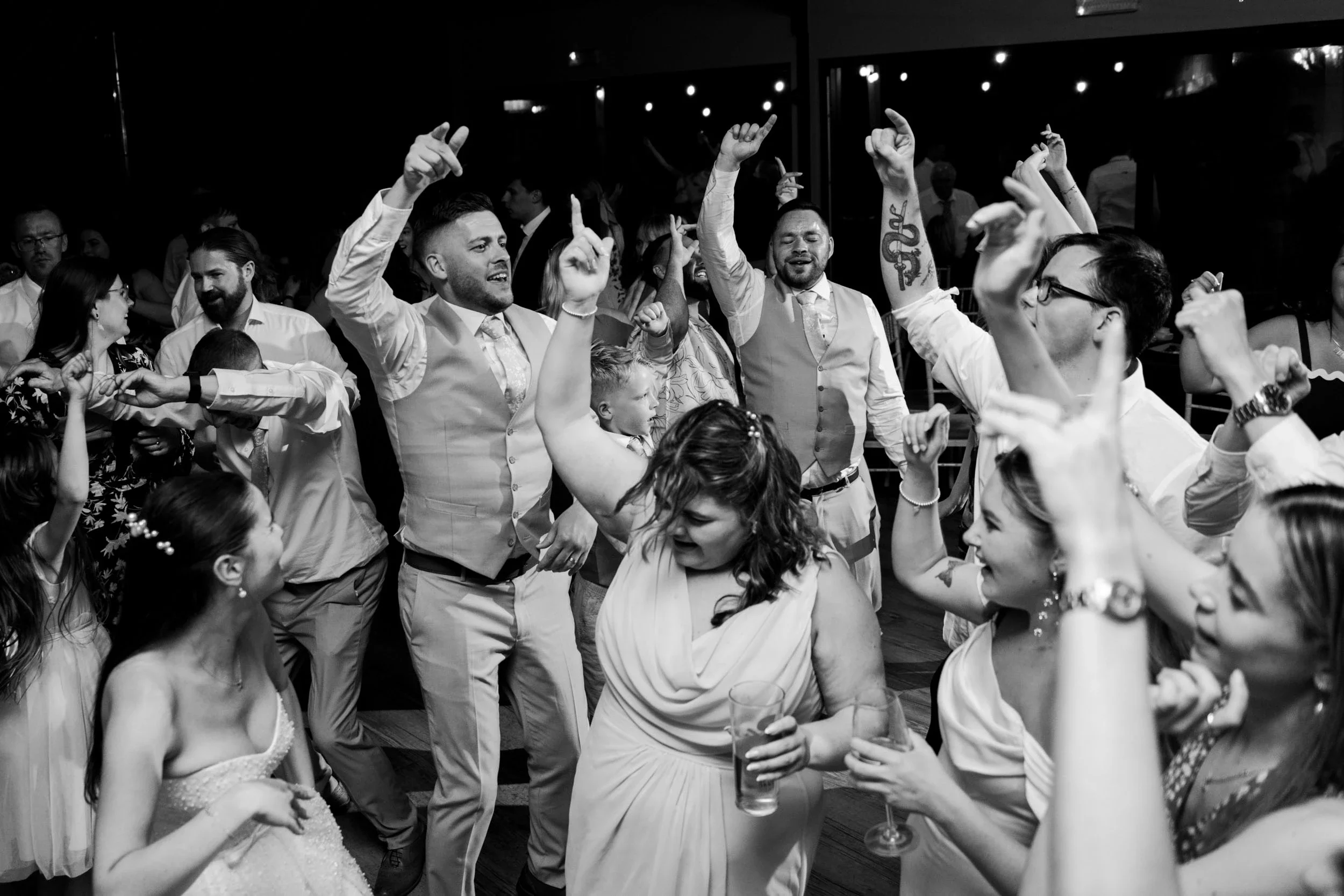 People dancing and celebrating at a wedding reception in a dimly lit venue, with smiles and raised hands.