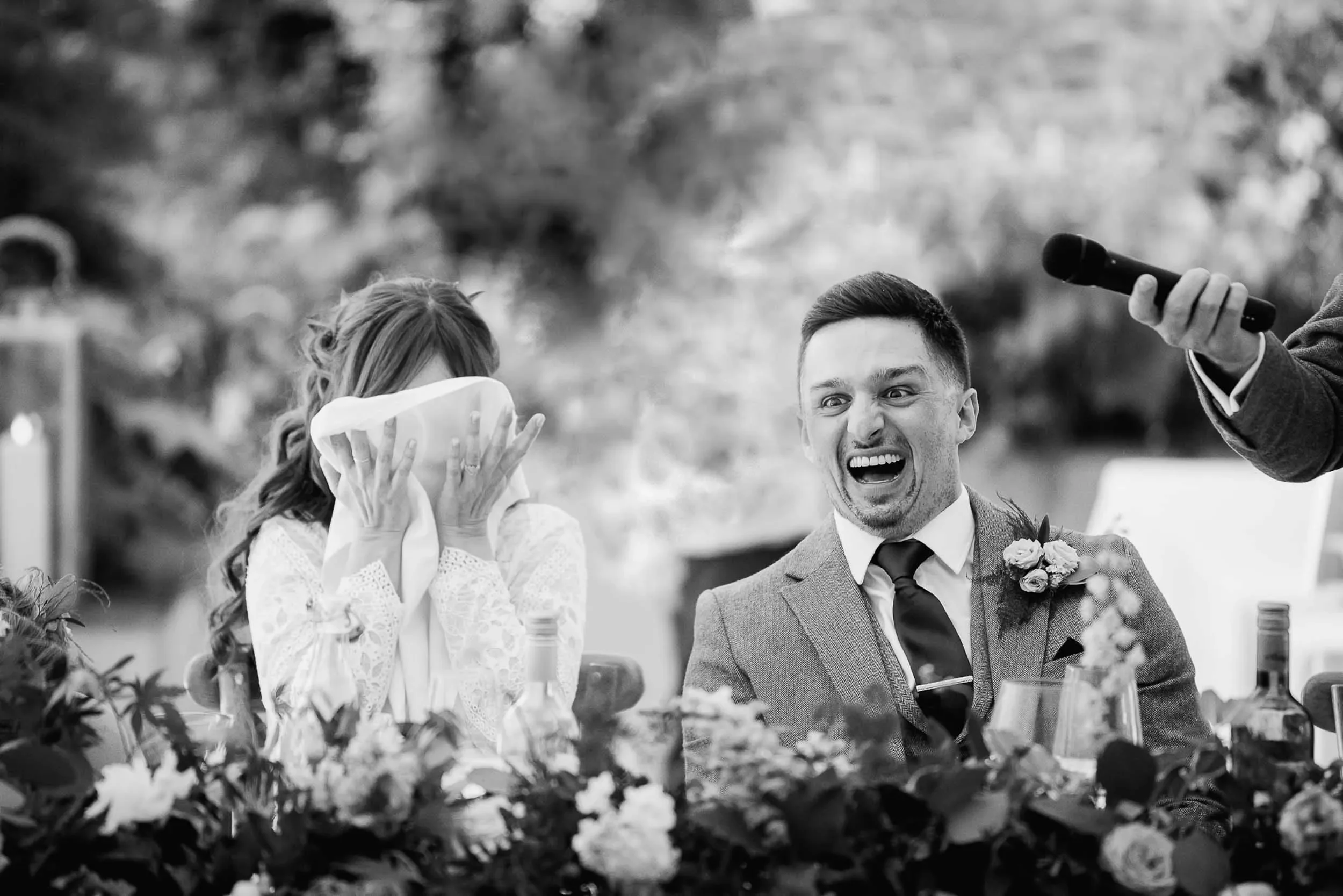 A woman covering her face with her hands and a man with a shocked or surprised expression at a wedding reception, with a microphone held toward the man.