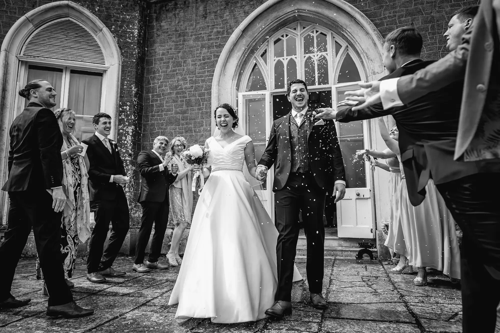 A bride and groom walking hand in hand outside a church, surrounded by friends and family throwing confetti, celebrating their wedding in black and white.