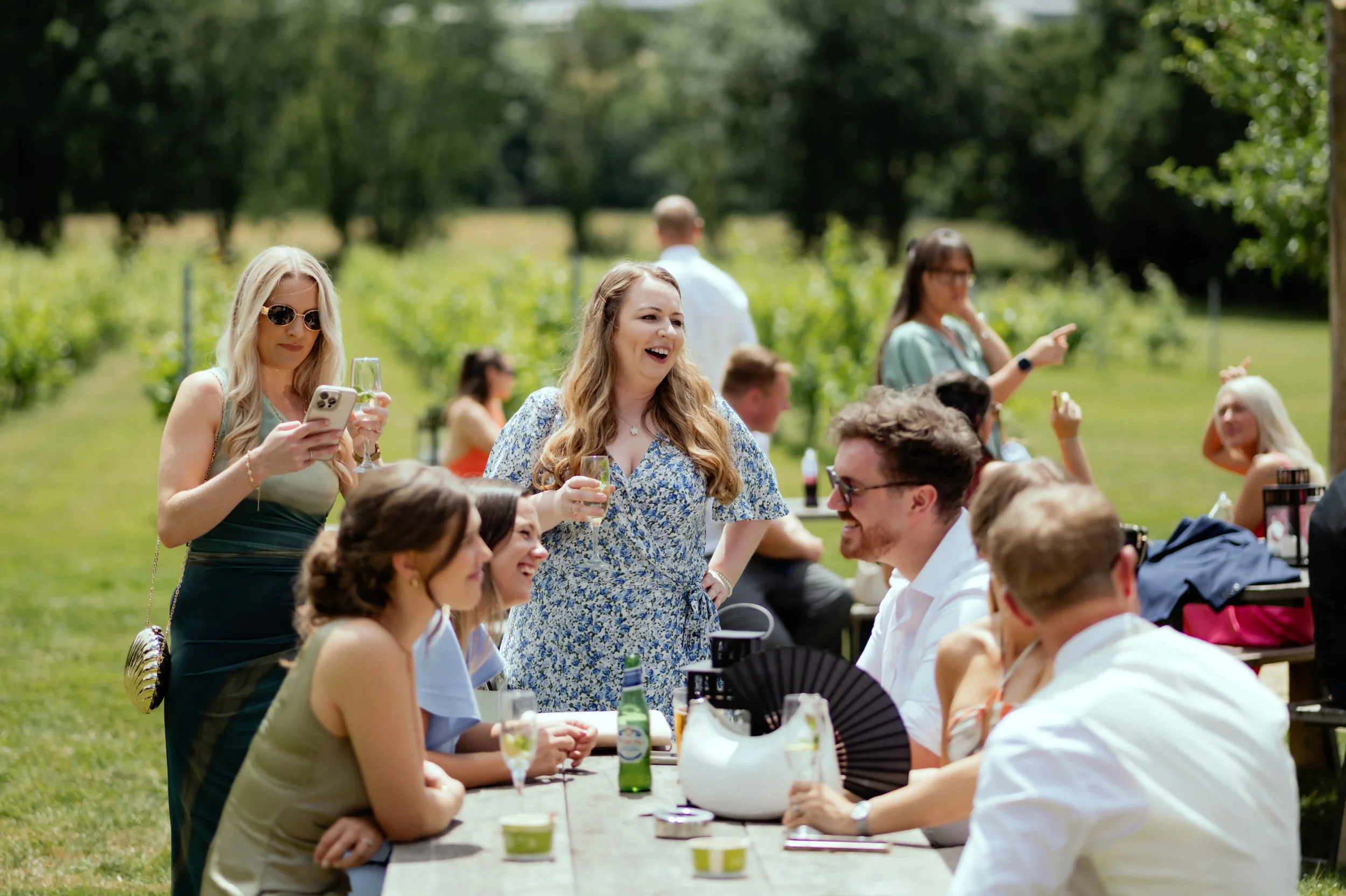 People at an outdoor gathering or party on a sunny day, sitting and standing near a long table in a green backyard or park area.