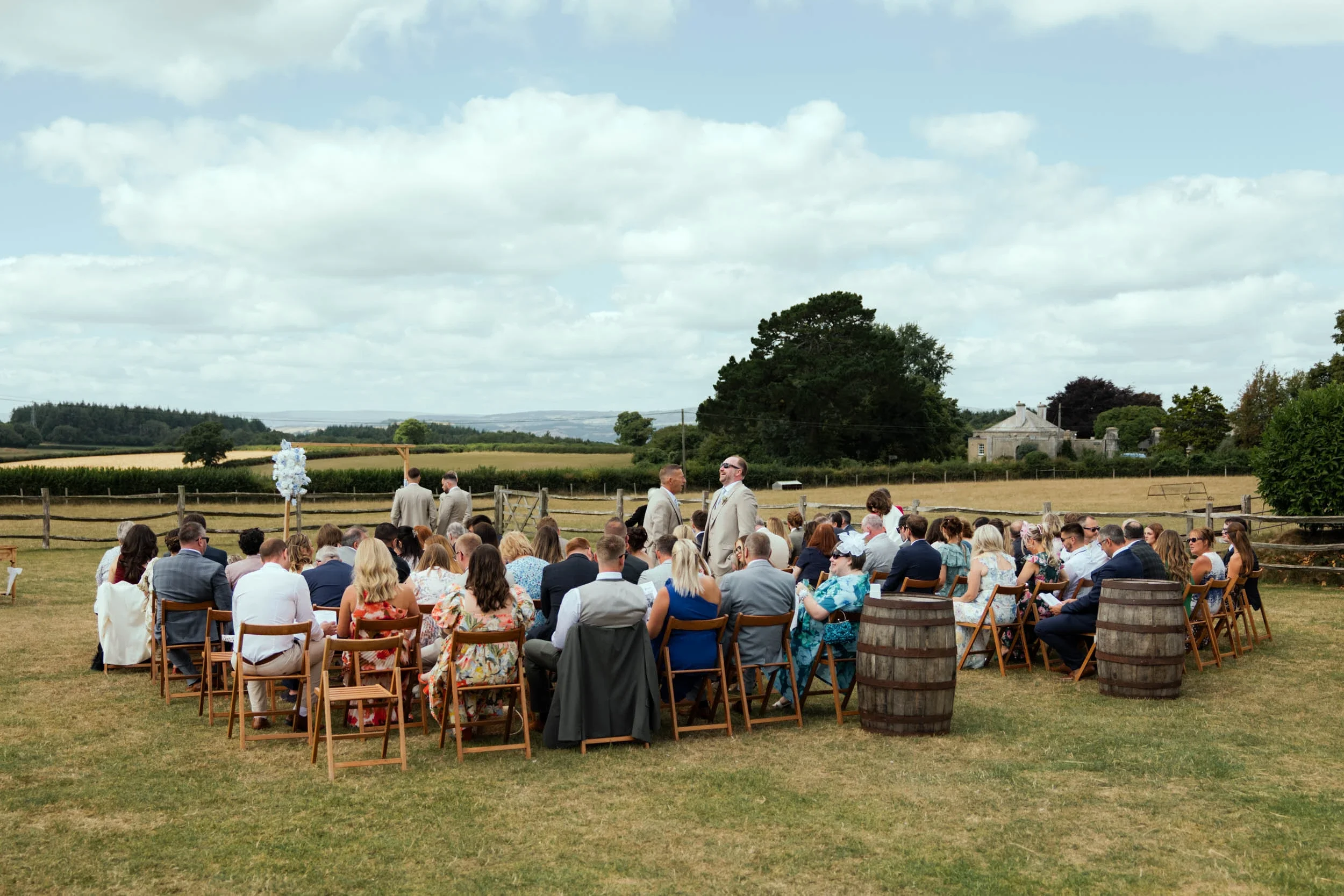Outdoor wedding ceremony with guests seated on wooden chairs on a grassy field, officiants in front, scenic countryside background with trees and a blue sky.