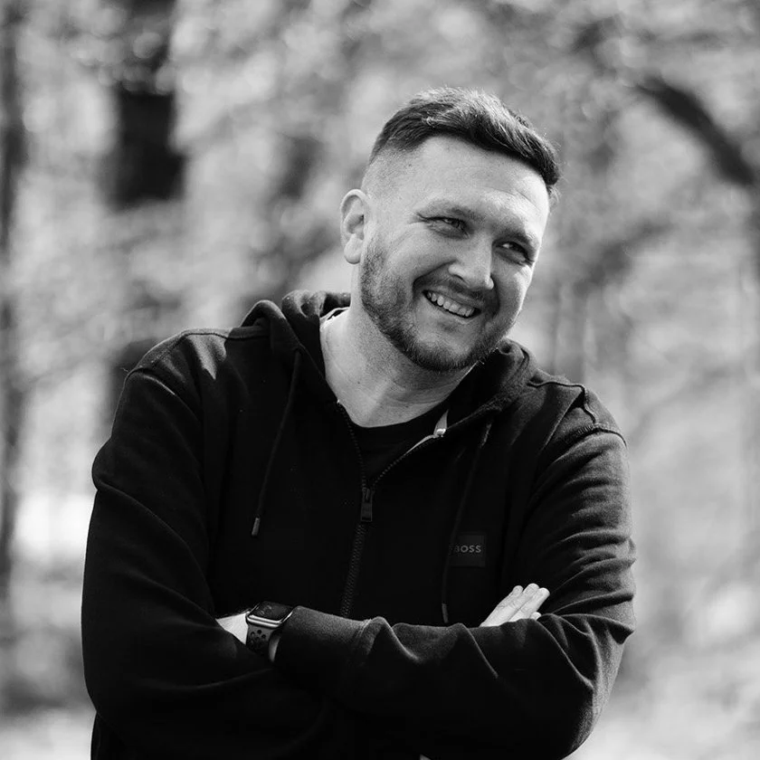 A smiling man with short hair and a beard, wearing a black hoodie, standing outdoors with trees in the background.