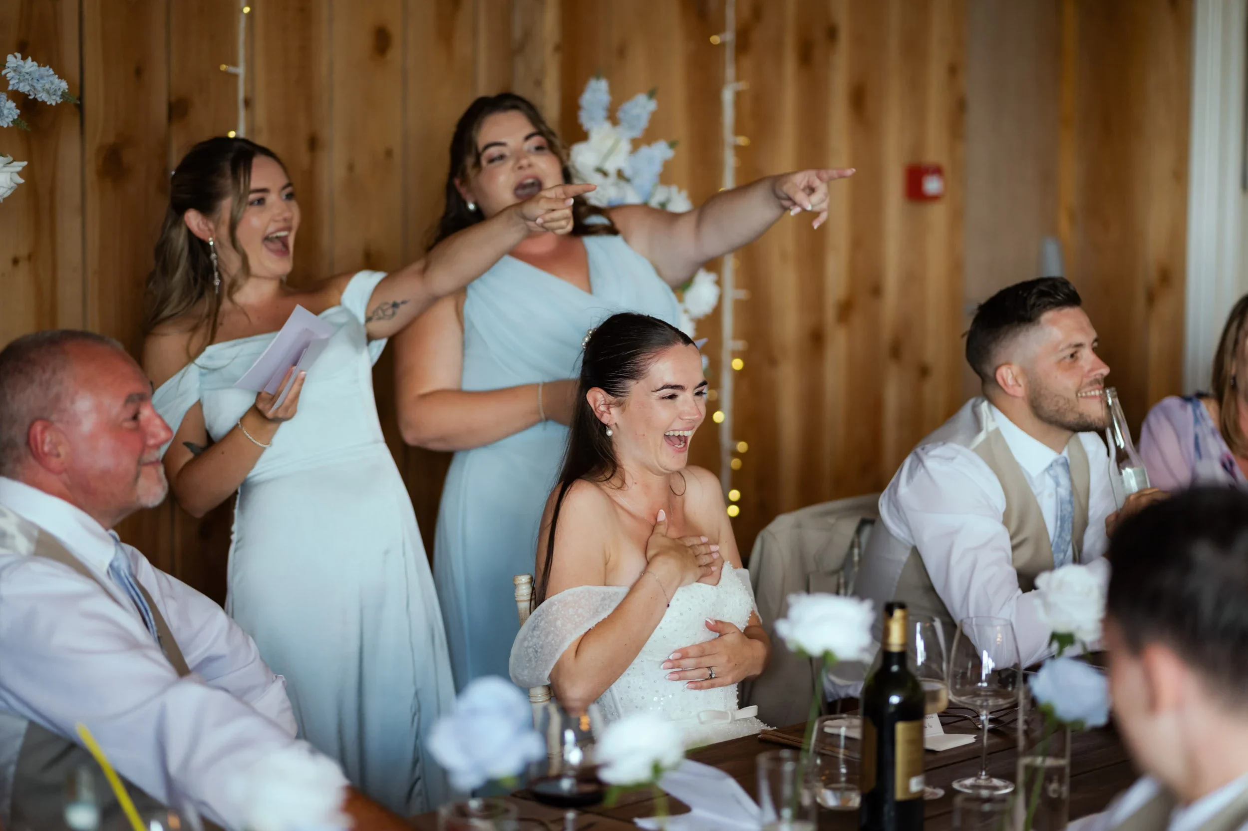 Group of people at a wedding reception, with women in dresses and men in shirts, smiling, laughing, and pointing, with a wooden wall background and floral decorations.